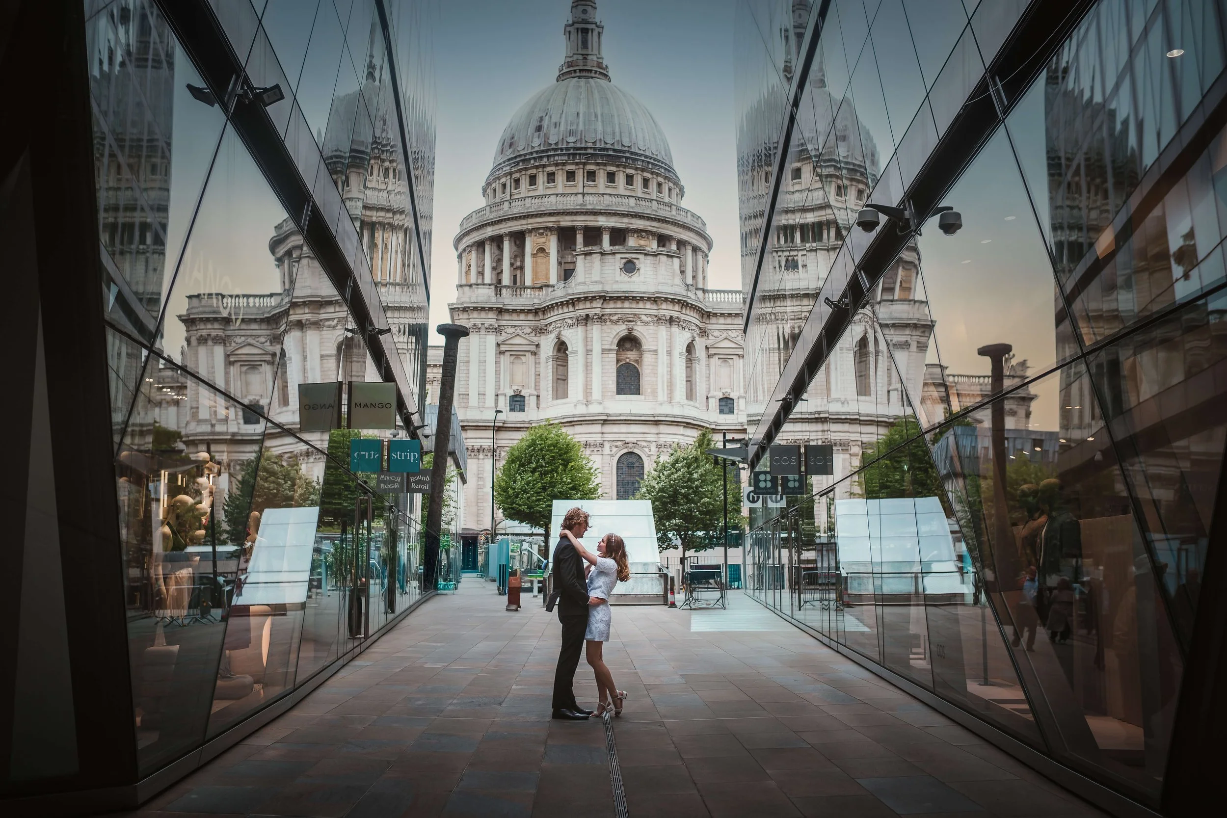 A couple is standing close and looking at each other in a modern glass corridor with reflections, with St. Paul's Cathedral visible in the background.