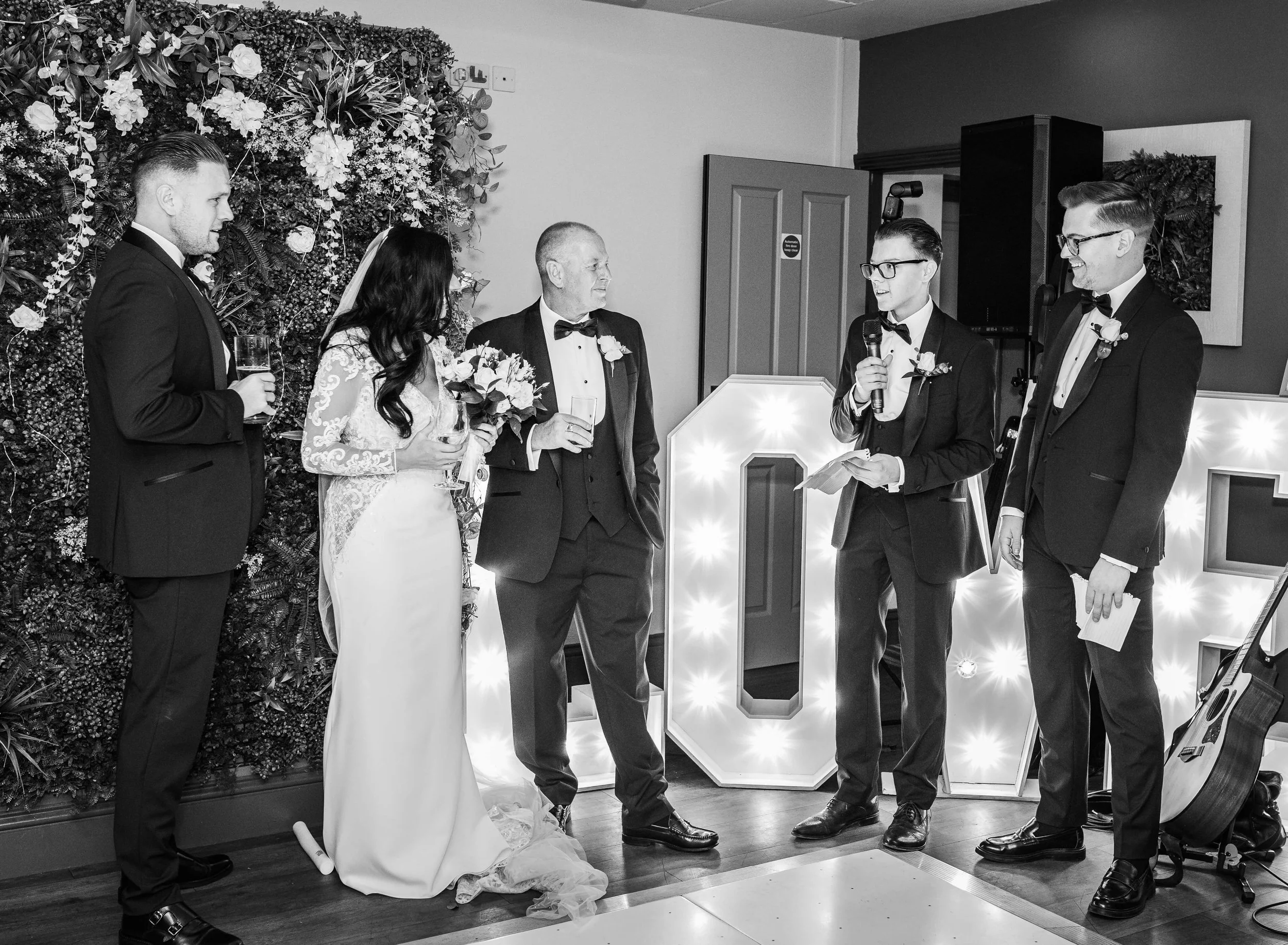 A black-and-white photo of a wedding ceremony with five people, including a bride, groom, and three men in suits. The bride, holding a bouquet, stands next to the groom and the man on the left. The woman is speaking into a microphone held by another 