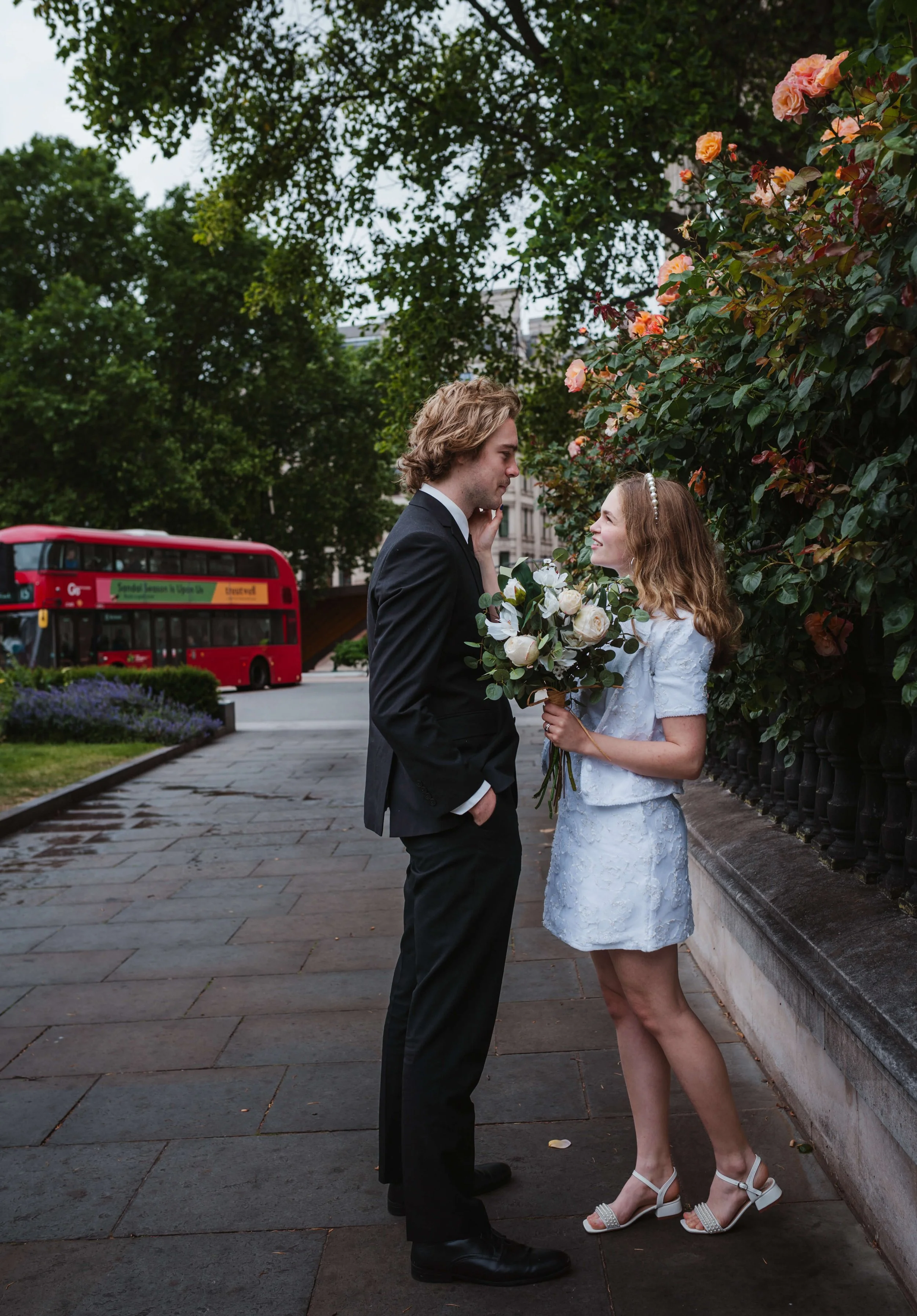 A young couple dressed in wedding attire standing on a city sidewalk, the woman holding a bouquet of white roses, looking into each other's eyes, with a red double-decker bus in the background and lush green trees.