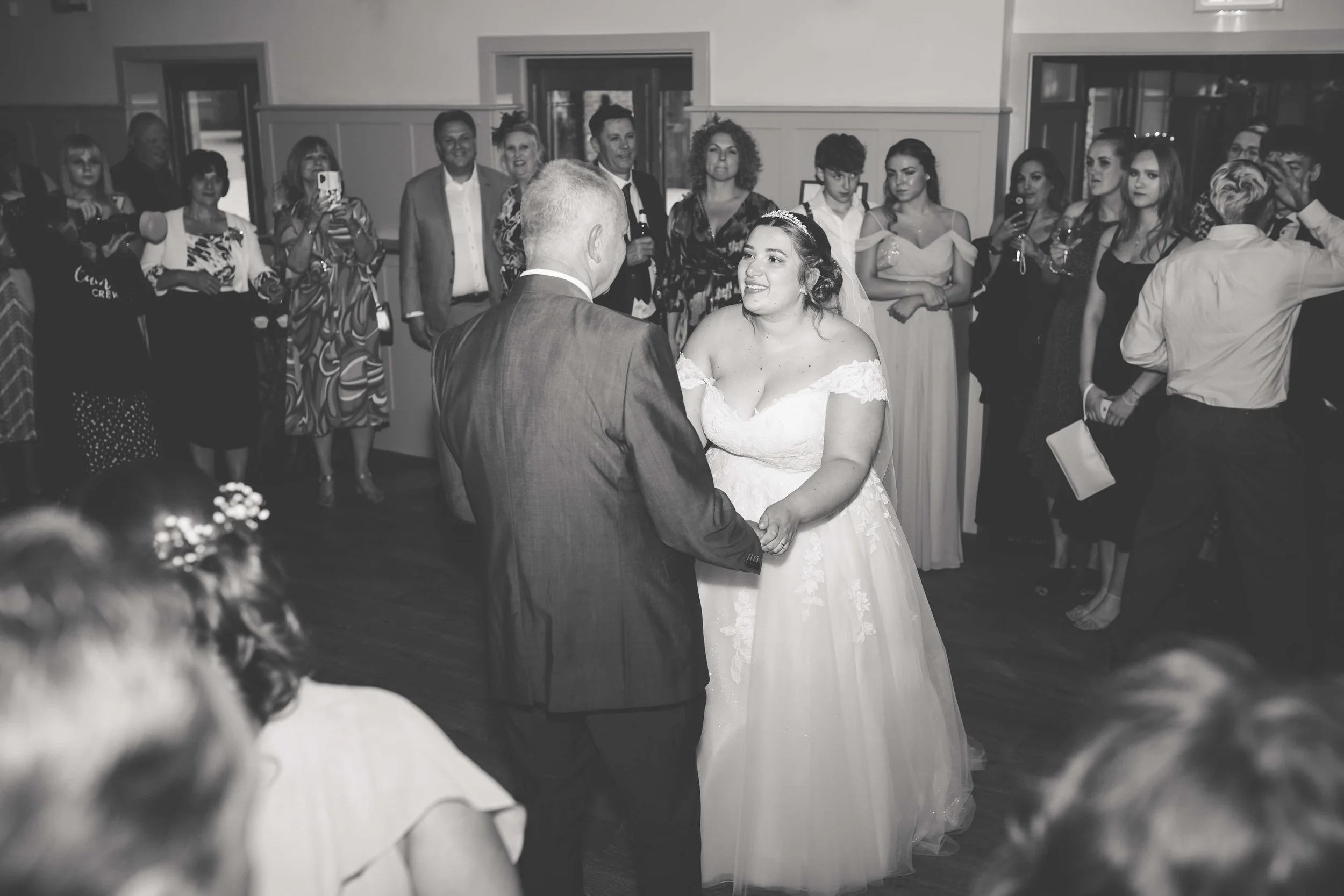 A bride and groom holding hands and dancing at their wedding reception, surrounded by seated and standing guests observing the moment, in a decorated indoor venue.