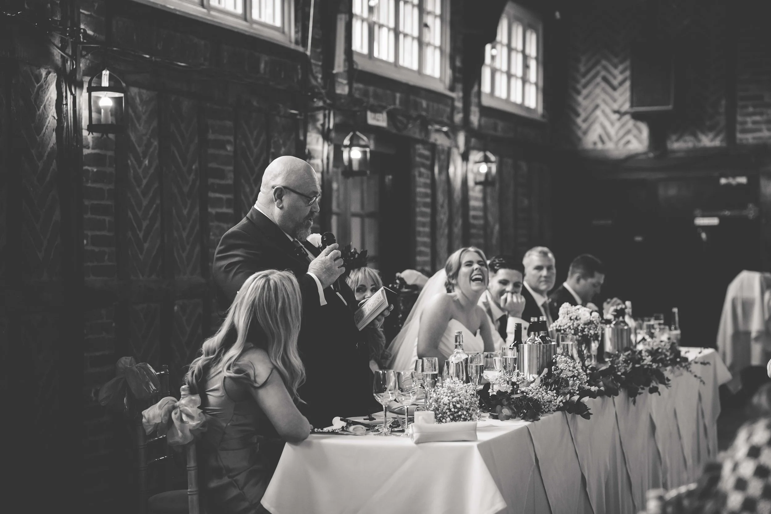 A wedding reception with a man giving a speech at a decorated head table. The bride is smiling and laughing, surrounded by groomsmen and bridesmaids, in a rustic venue with wood walls and large windows.