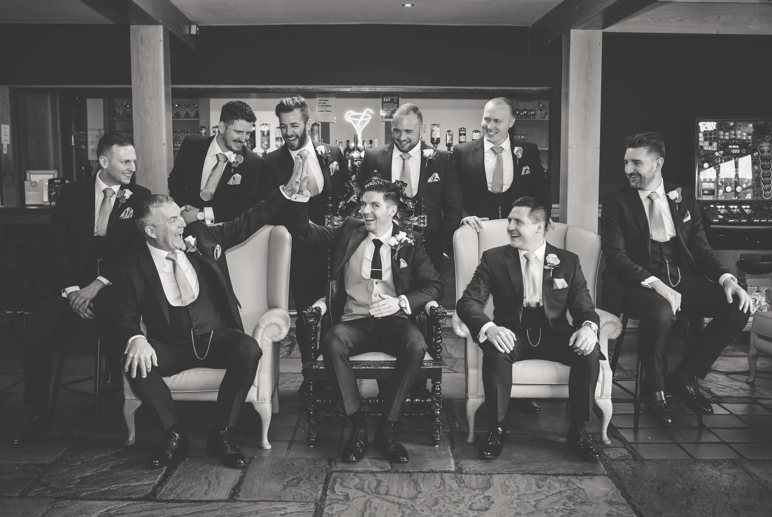 A group of men dressed in suits with boutonnières, sitting and standing in a lounge area, celebrating with high-fives and smiles.