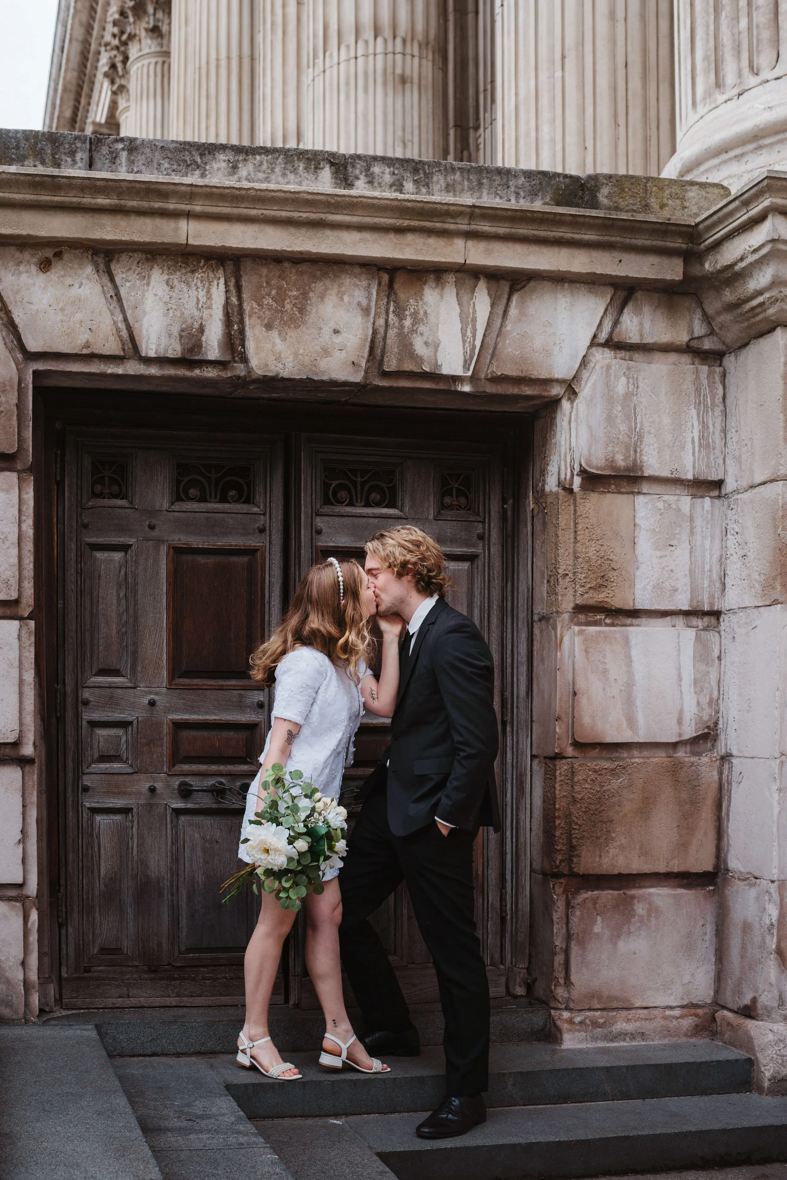 A couple dressed in wedding attire sharing a kiss in front of a large old wooden door with stone architecture.