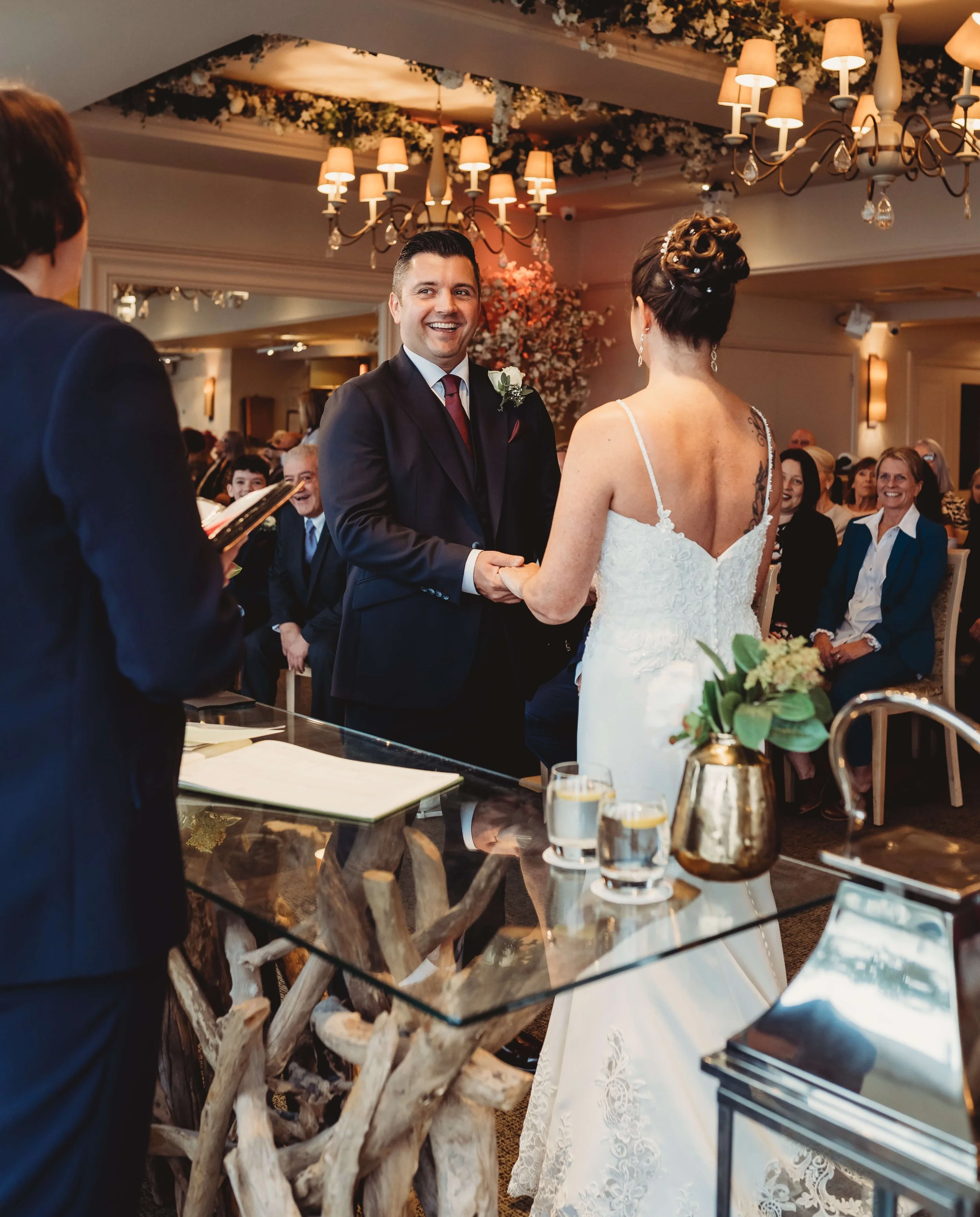 Bride and groom exchanging vows during wedding ceremony in a decorated indoor venue with chandeliers and floral arrangements.