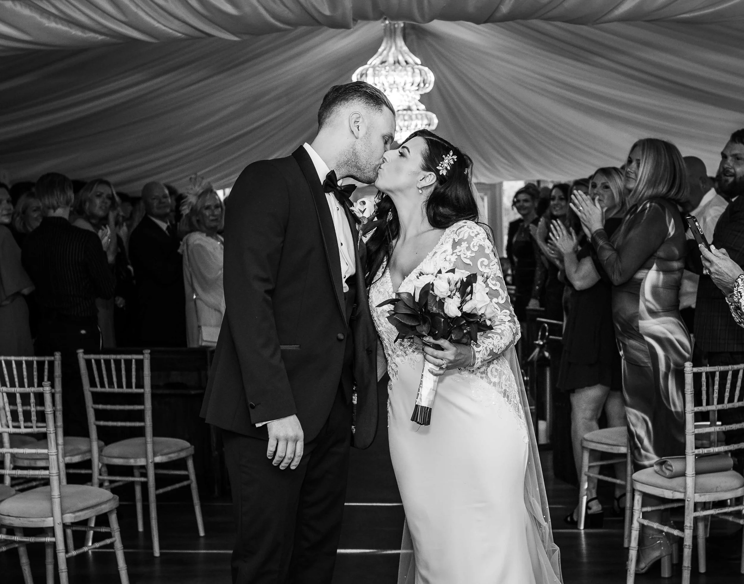 A black and white photo of a wedding ceremony with a bride and groom sharing a kiss in the center, surrounded by seated and standing guests in a decorated tent.