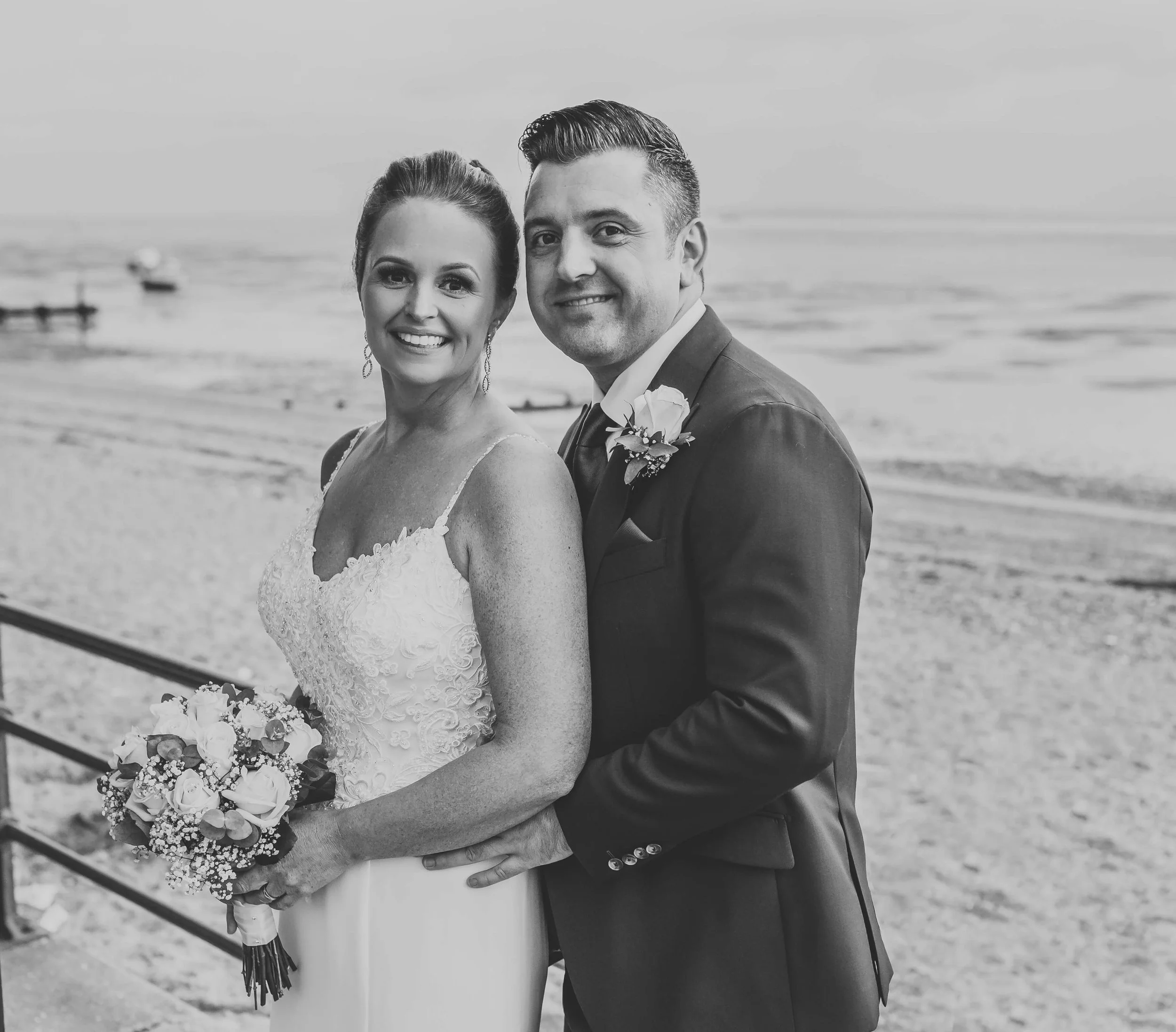 Black and white photo of a bride and groom standing on a beach, smiling at the camera. The bride wears a lace wedding dress and holds a bouquet, while the groom is in a suit with a boutonnière.