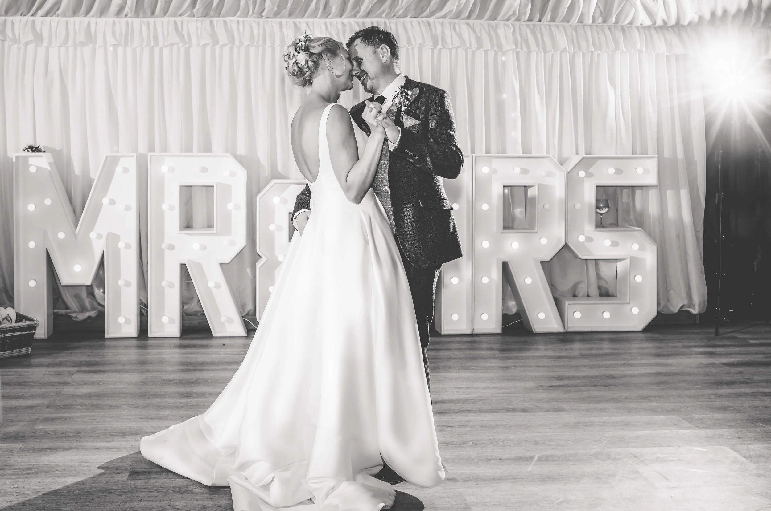 A bride and groom share a dance at their wedding reception, with large illuminated letters spelling 'MRS' in the background.