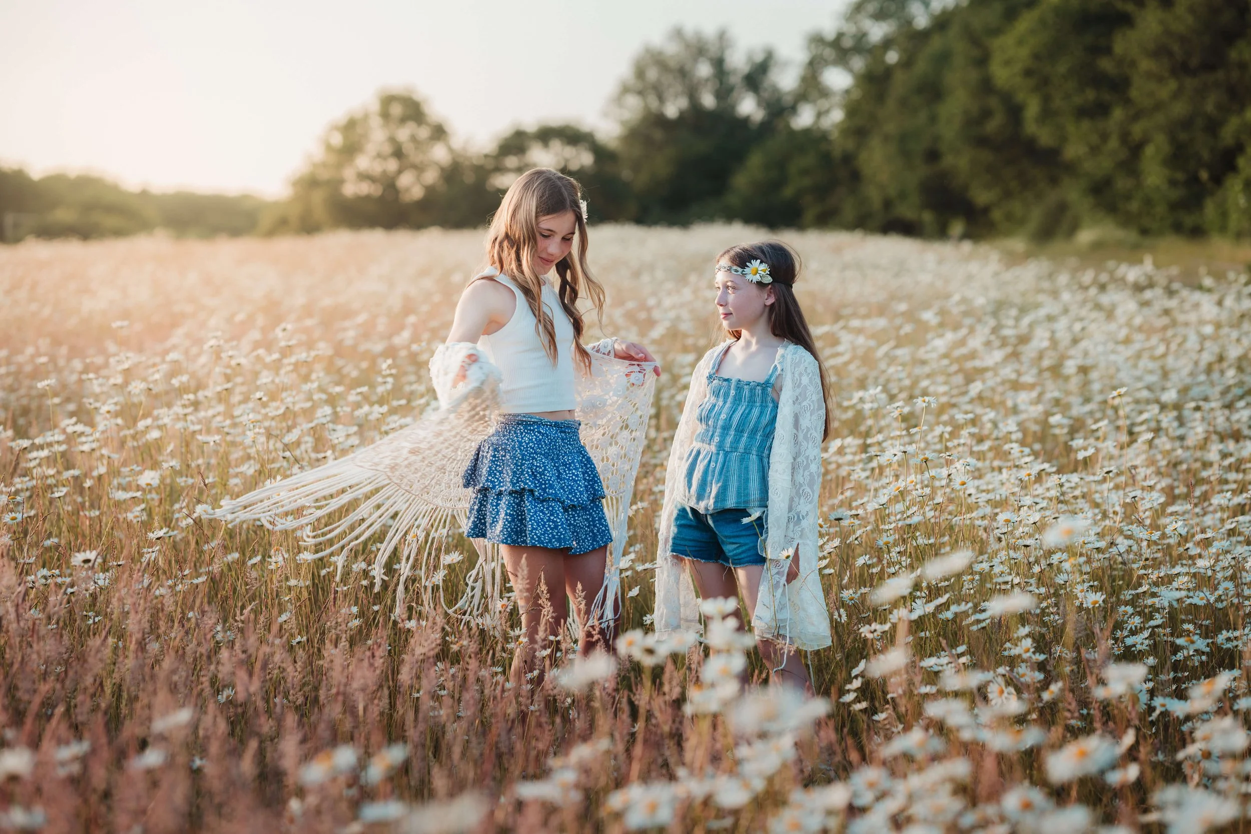 Two young girls standing in a field of daisies during sunset, one holding a parasol, both dressed in summer clothes and with floral accessories.