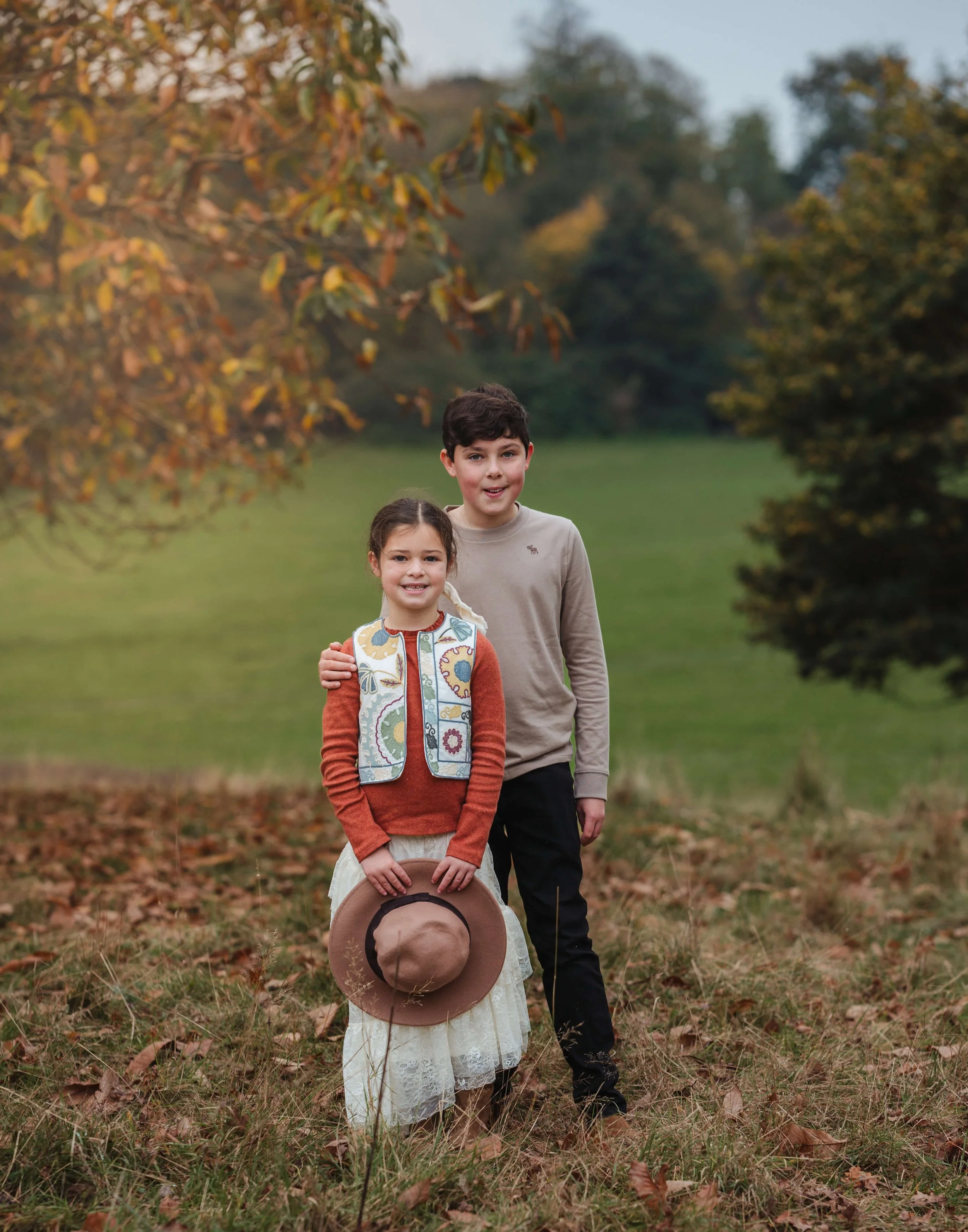 A young girl and boy standing outdoors in a field with trees and autumn leaves. The girl is holding a hat and smiling, while the boy stands beside her with his arm around her shoulder. It appears to be fall.