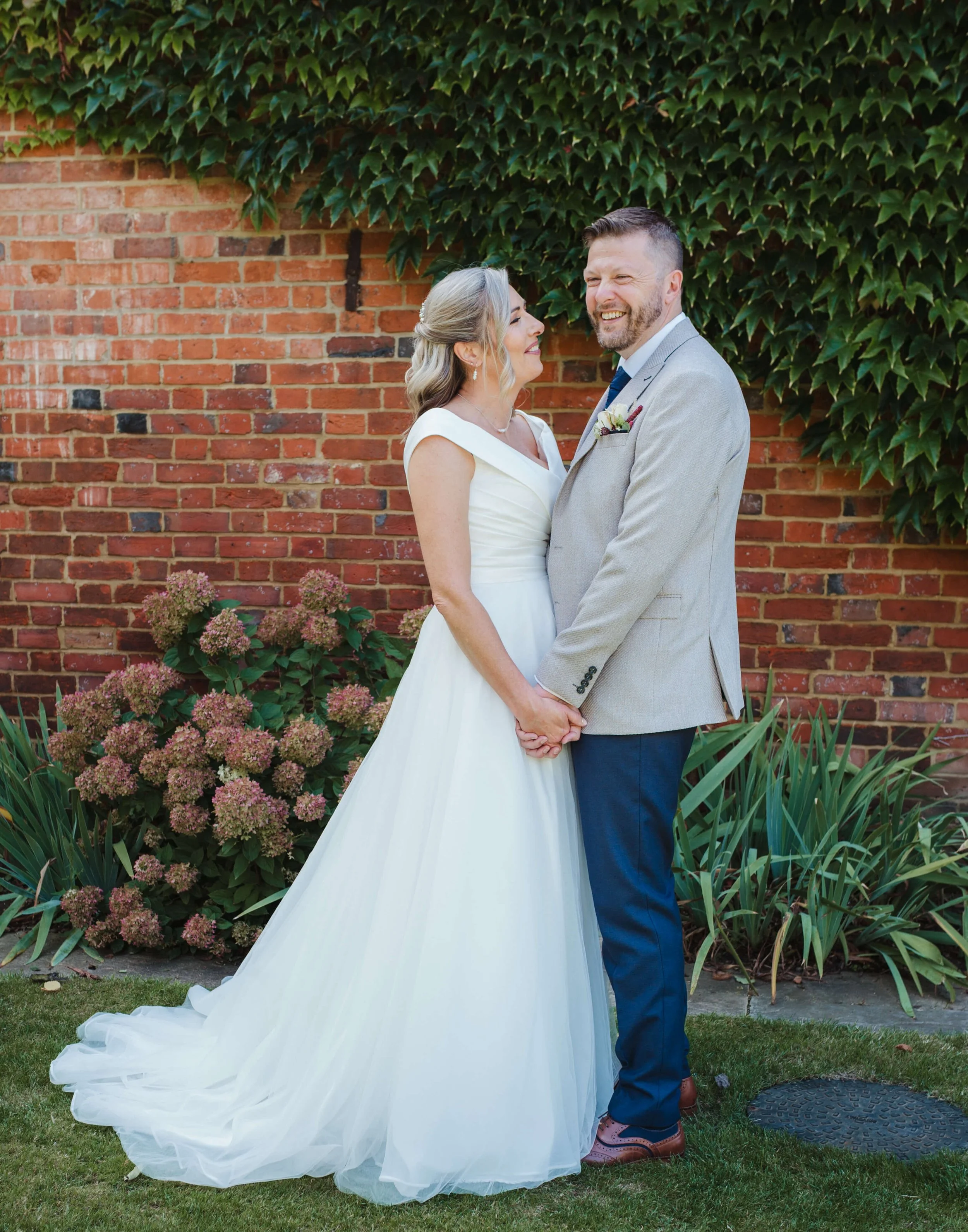 A bride and groom holding hands and smiling at each other outdoors with a brick wall, green ivy, and flowering plants in the background.