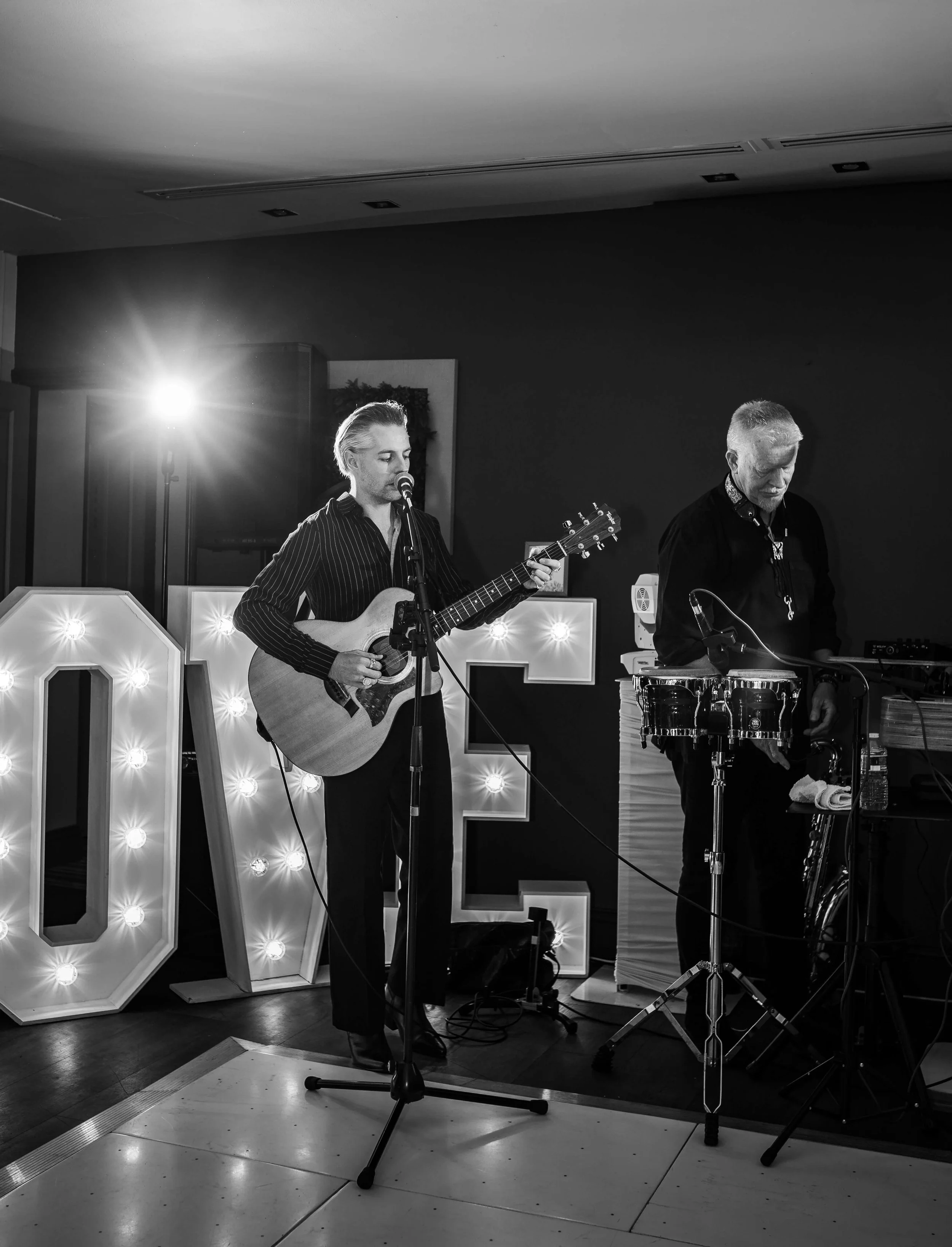 Black and white photo of two musicians performing on a stage with illuminated large letter signs.