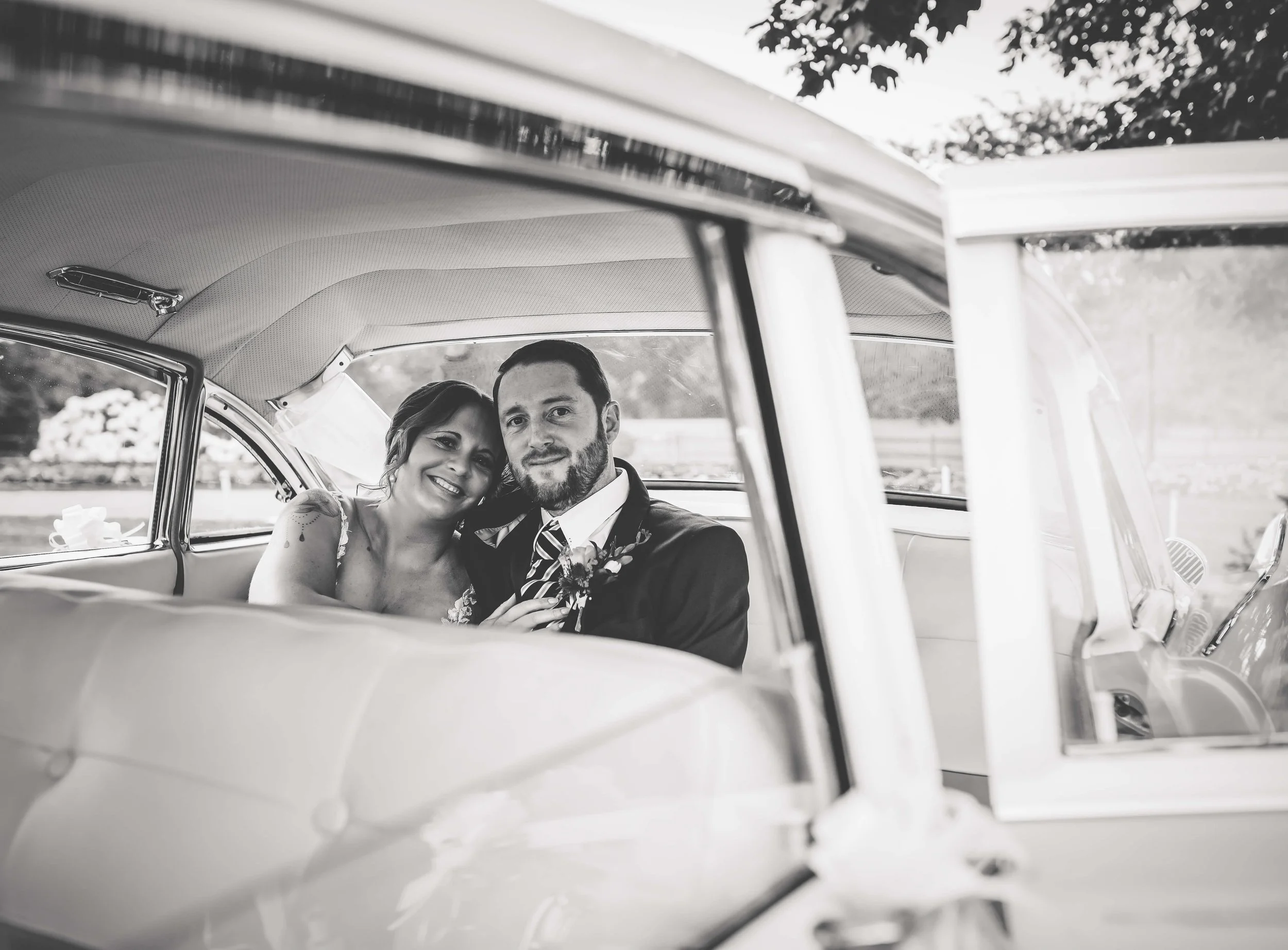 A black-and-white photo of a bride and groom sitting inside a vintage car, smiling and looking out of the window.