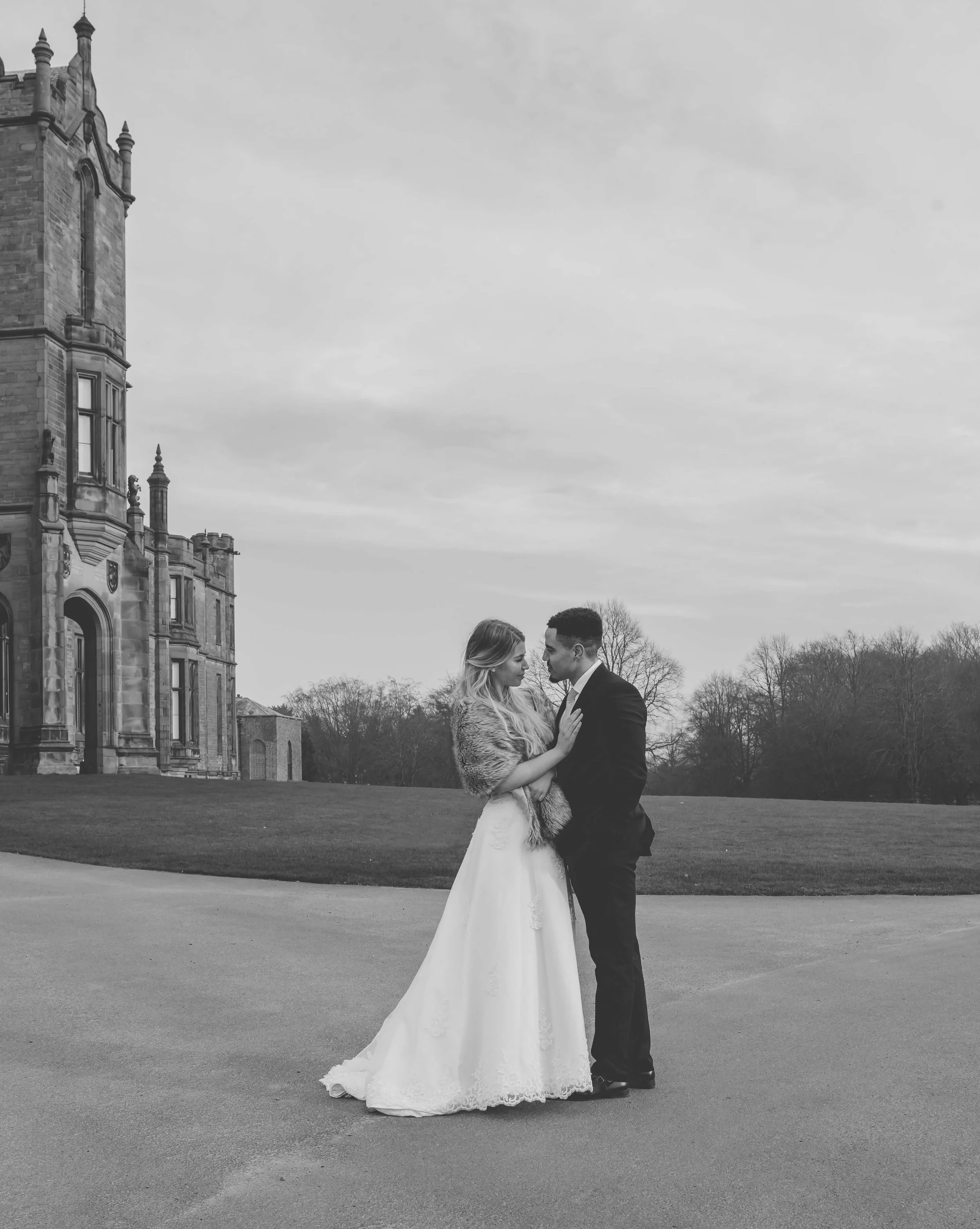 Black and white photo of a bride and groom standing close, facing each other with foreheads touching, outdoors near a historic castle-like building.