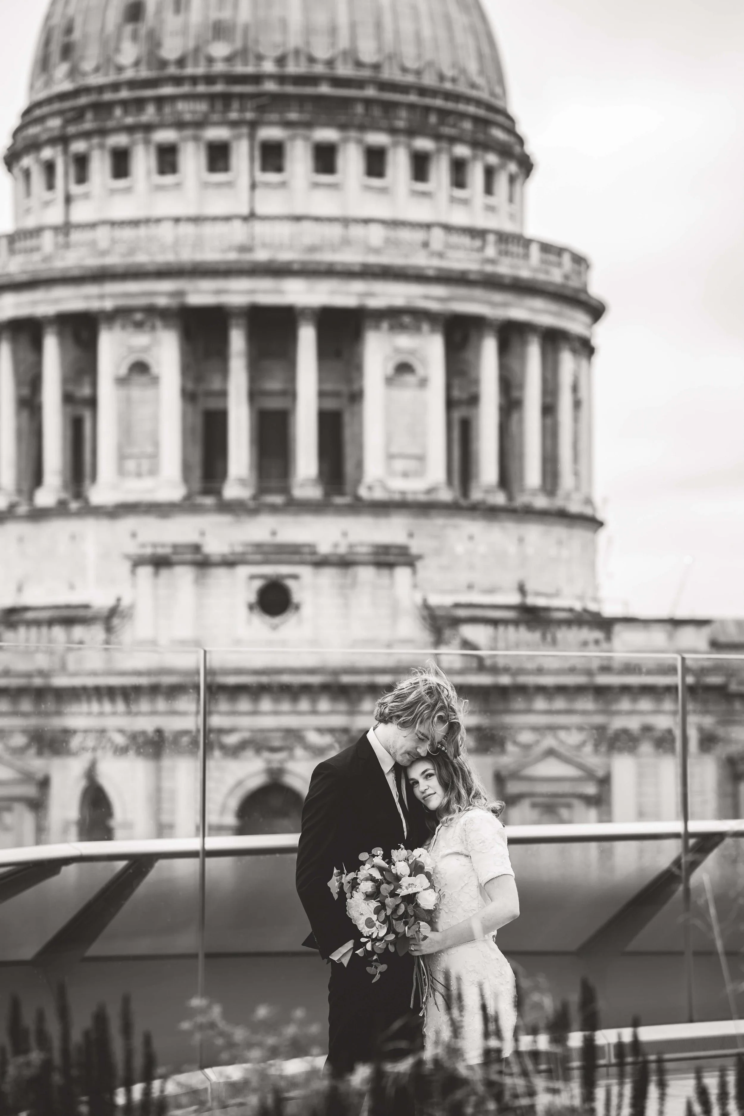 Black and white photo of a couple on a boat, with the United States Capitol building in the background. The man is in a dark suit and the woman is in a light dress, holding a bouquet of flowers.