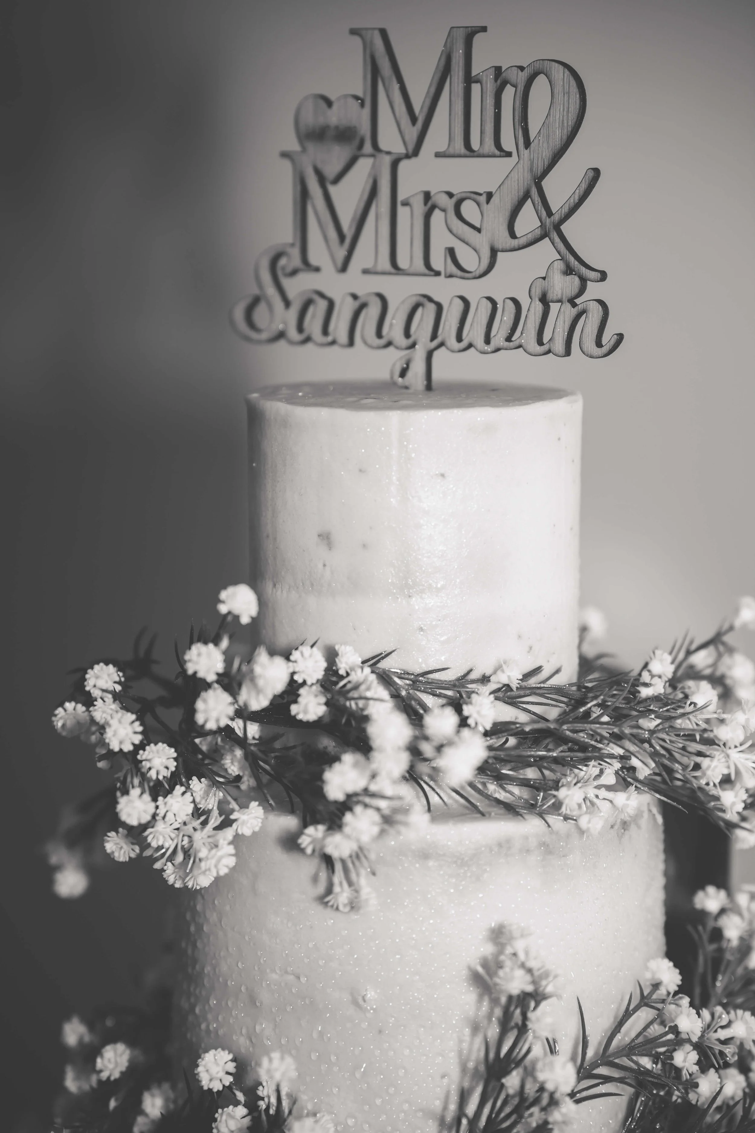 A wedding cake with white frosting, decorated with small white flowers and green foliage, topped with a "Mr & Mrs Sangwin" cake topper.