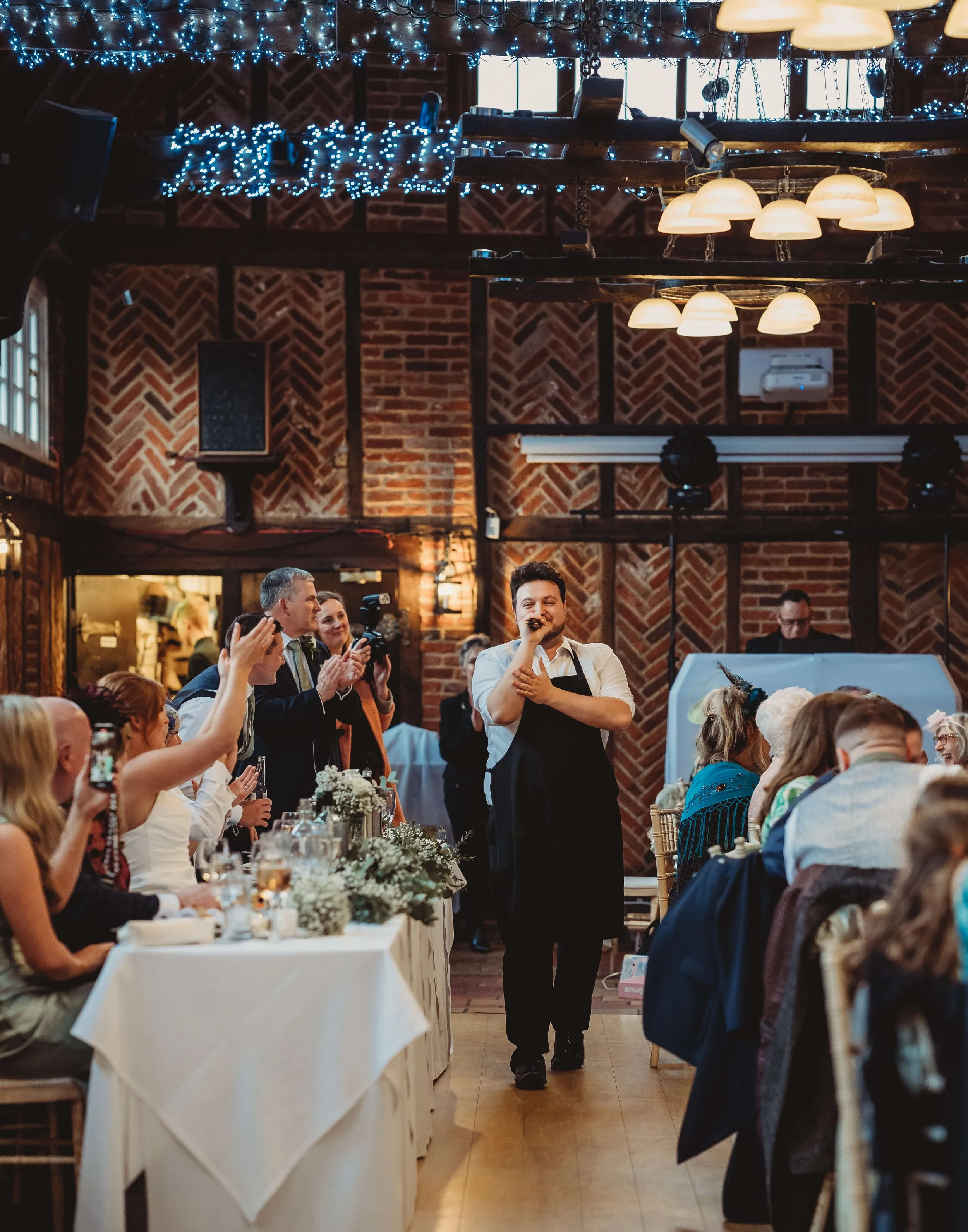 A man in a white shirt and black apron singing into a microphone at a wedding reception, surrounded by guests seated at long tables with floral decorations in a rustic venue with brick walls and string lights.