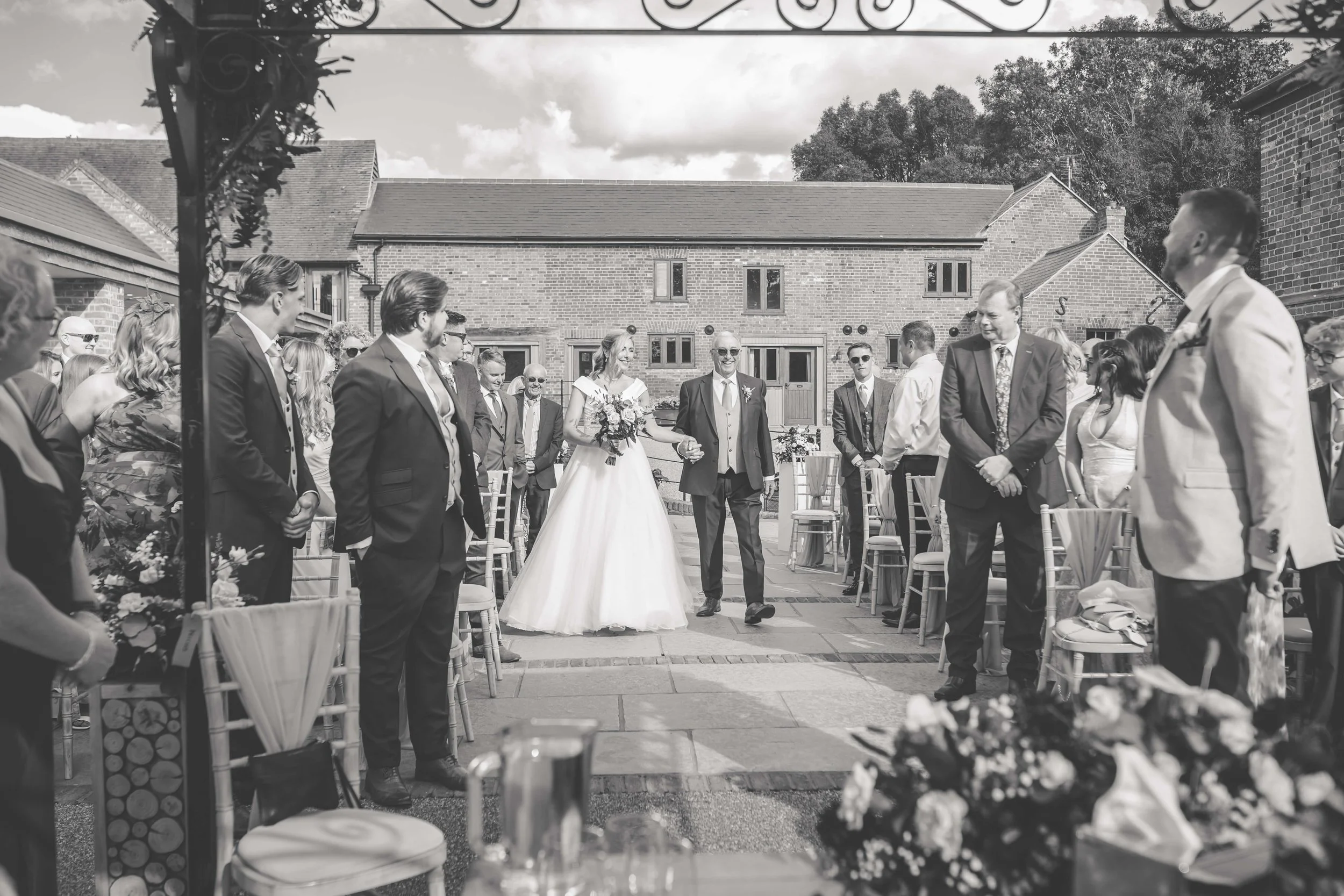 A black-and-white photo of a wedding ceremony outdoors in a courtyard. The bride, wearing a long white gown and holding a bouquet, walks down the aisle hand-in-hand with an older man, possibly her father. Guests are seated on both sides, standing and