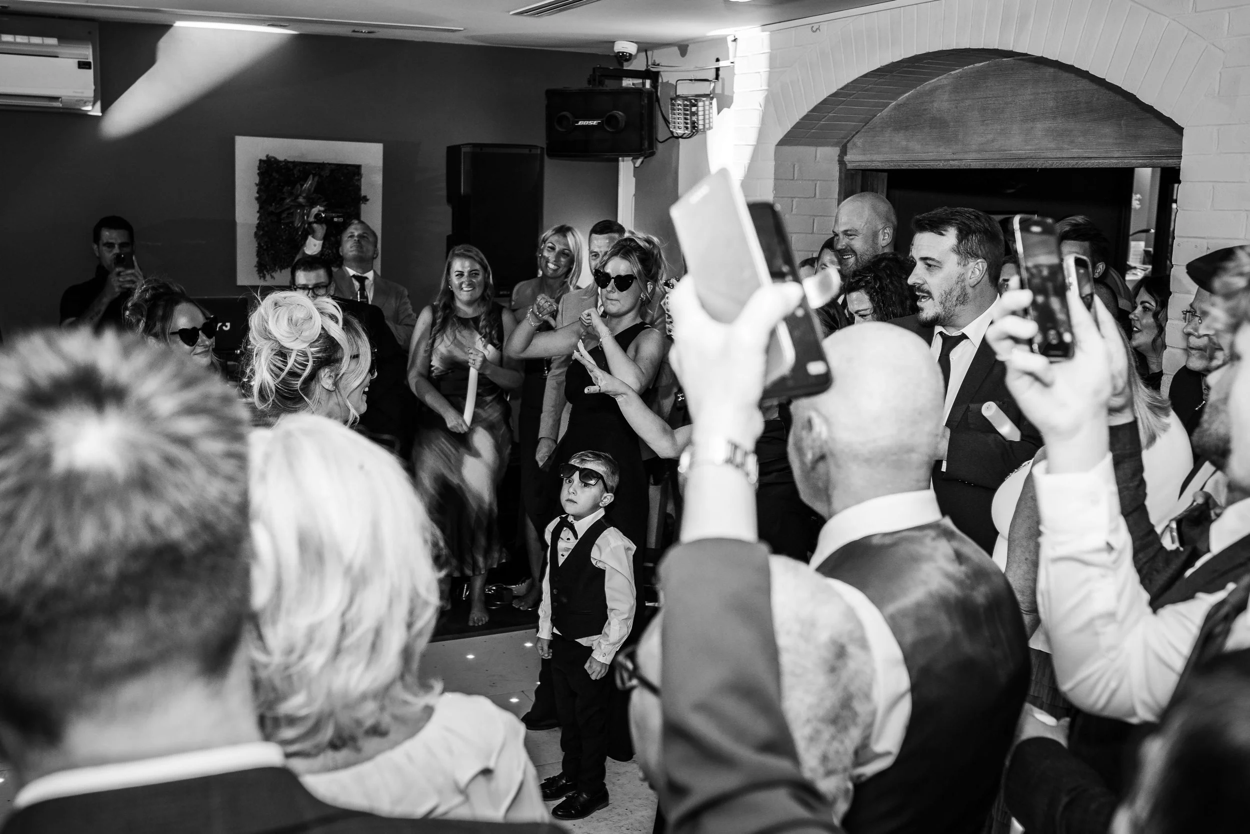 A black and white photo of a lively party or wedding celebration with many guests, some taking photos, and a young boy in formal attire standing in the center.
