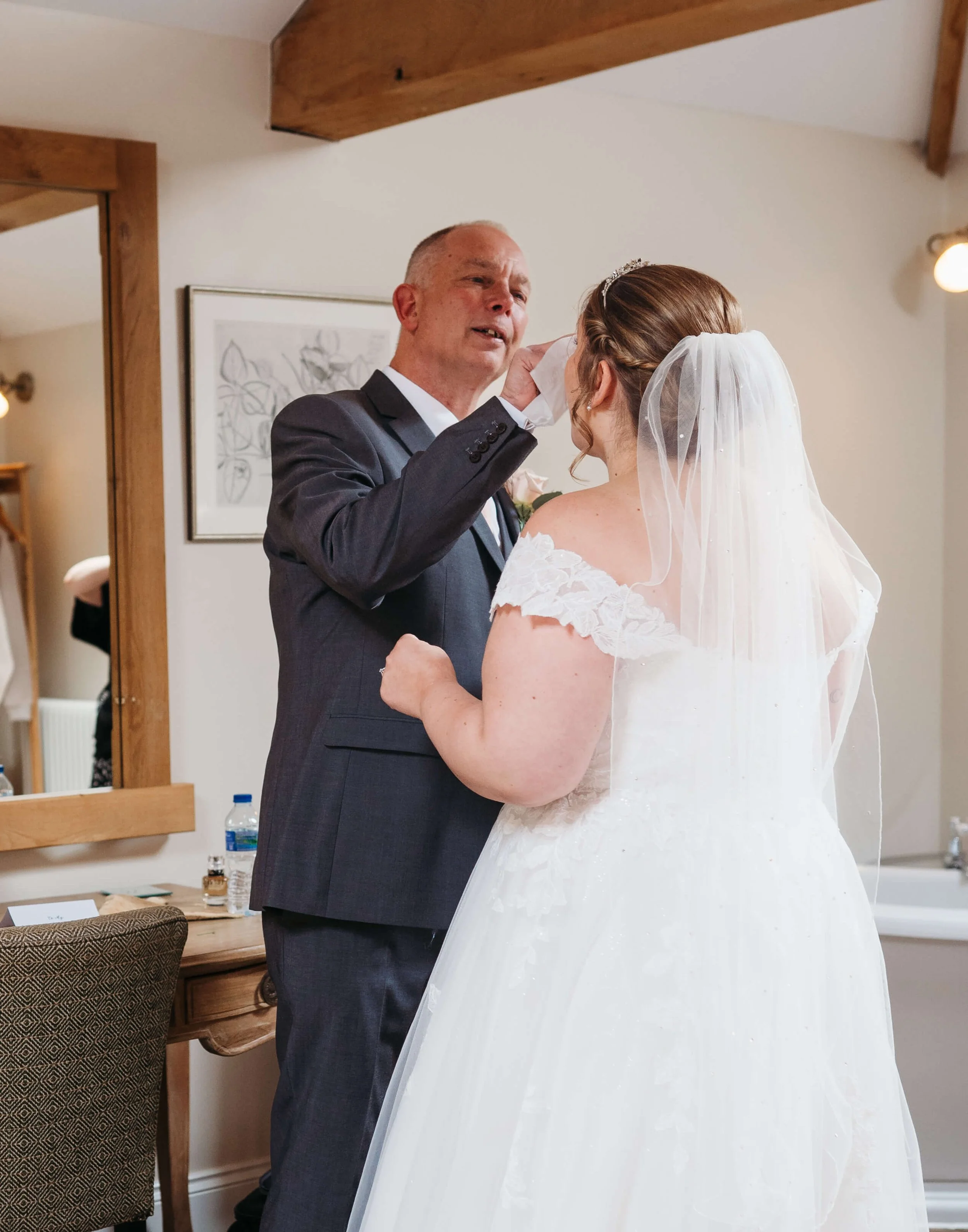 A bride in a white wedding gown and veil tears up while a man in a suit comforts her in a cozy room.