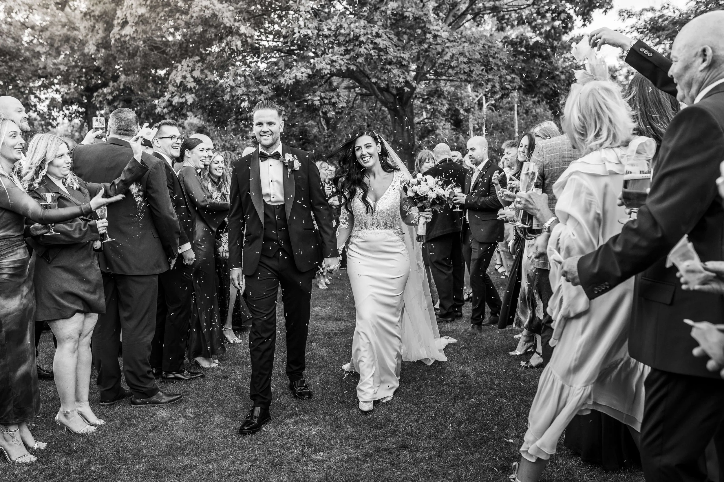 Black and white photo of a newlywed couple walking hand-in-hand through a crowd of guests outdoors, with guests celebrating and taking photos, trees in the background.