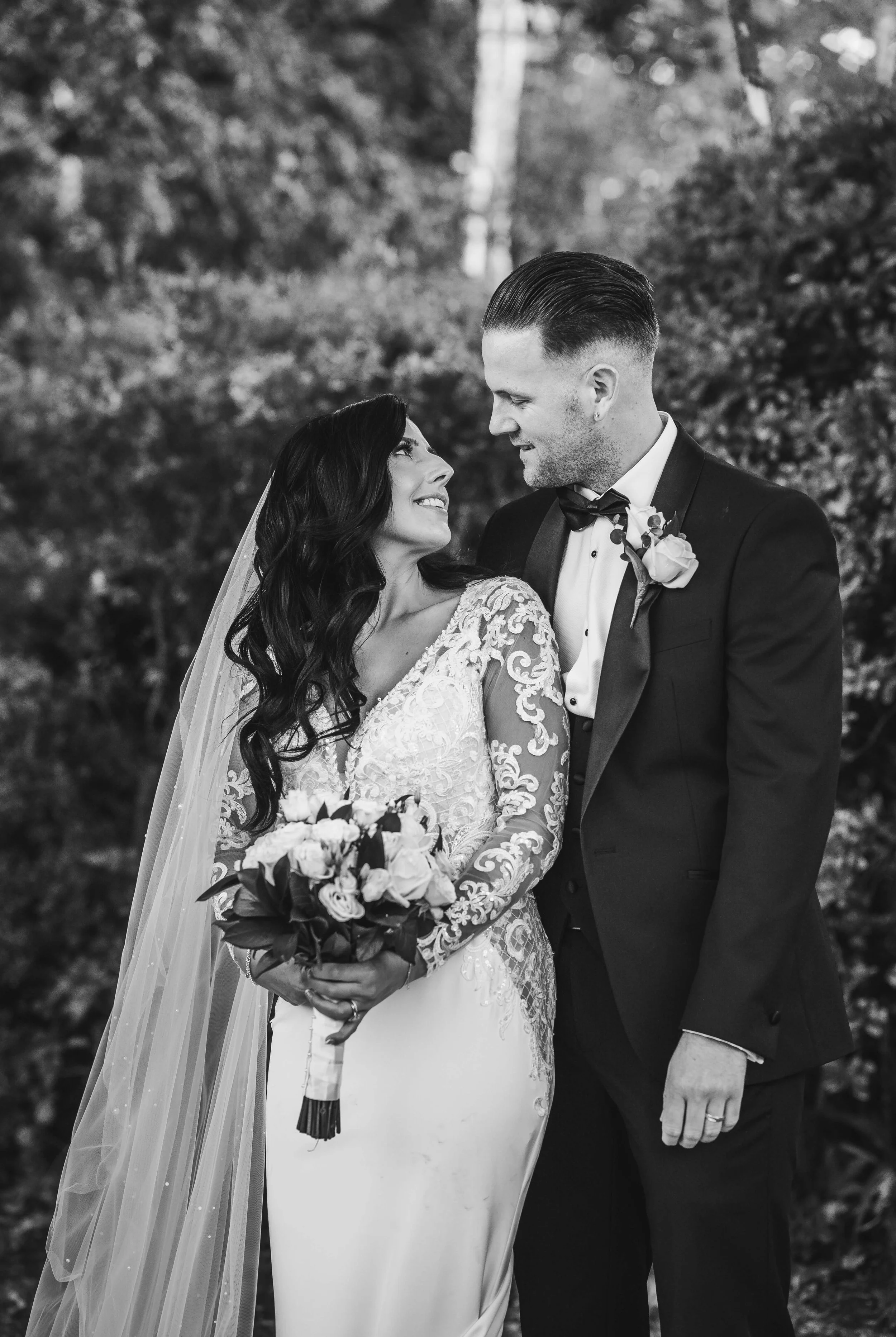 Black and white photo of a bride and groom looking at each other, standing outdoors with trees in the background. The bride is holding a bouquet and wearing a lace wedding dress, while the groom is dressed in a tuxedo with a boutonniere.