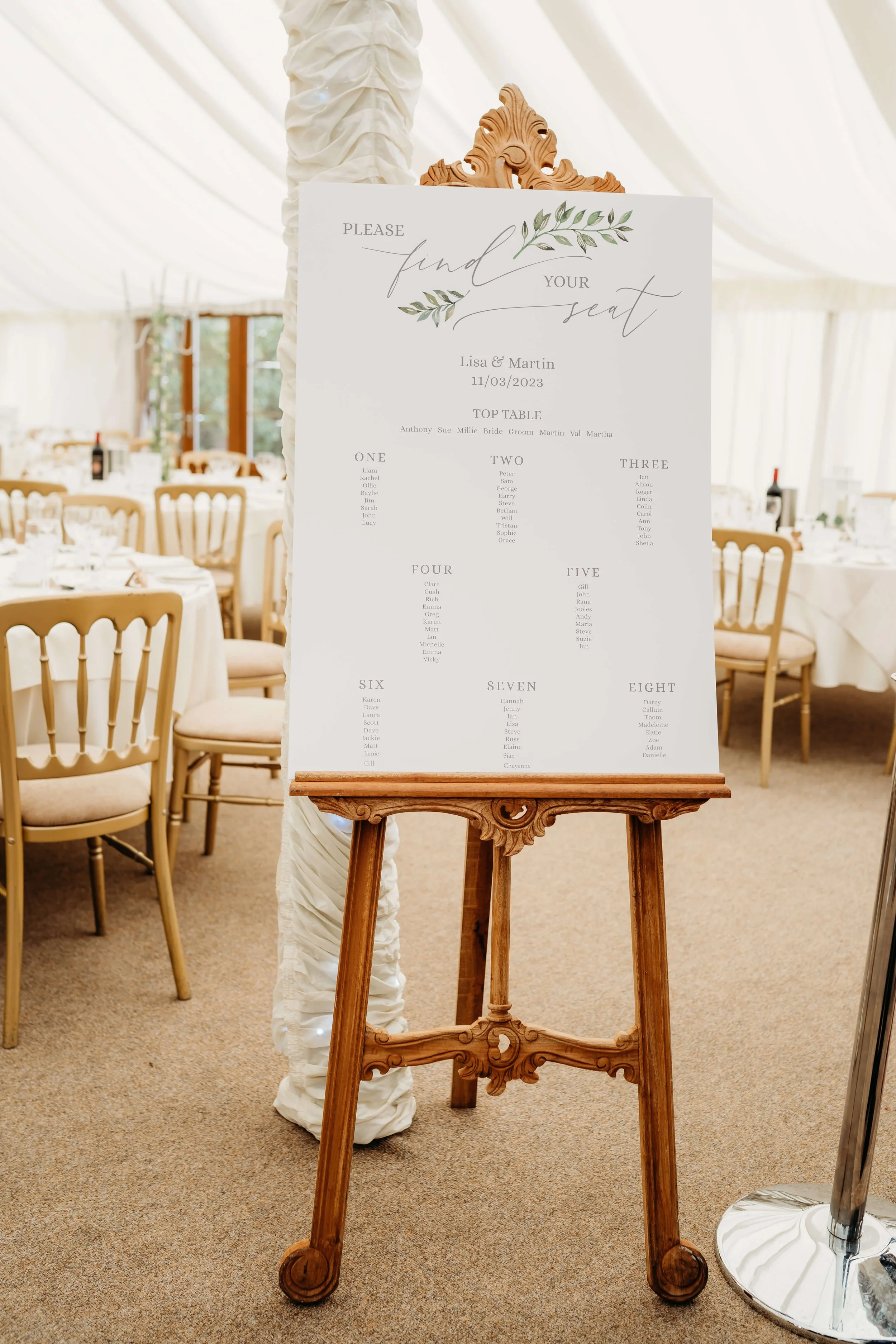 A wedding seating chart displayed on an ornate wooden easel in a decorated banquet hall with round tables and chairs, and a white draped ceiling.