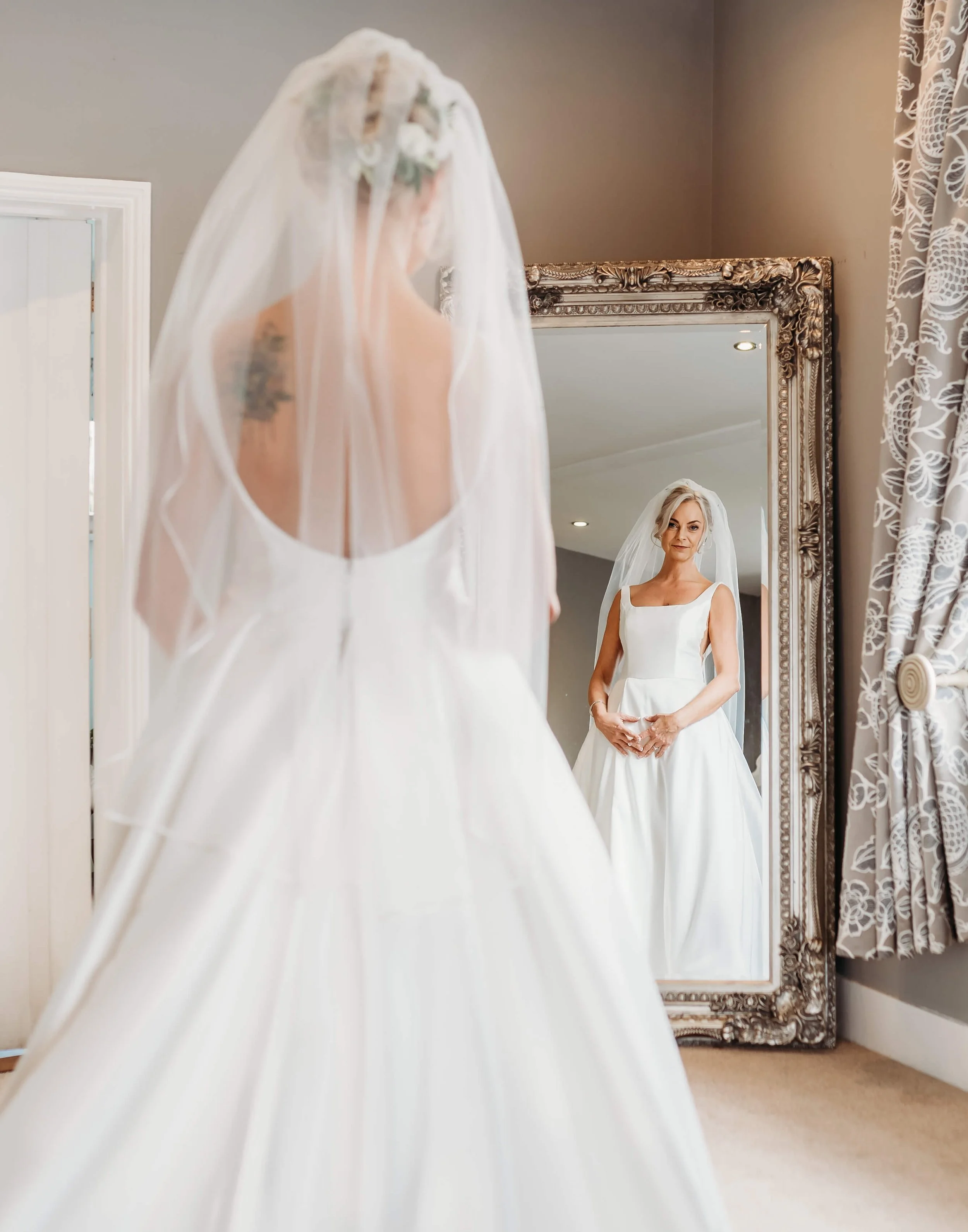 A bride in a wedding dress and veil looking at herself in a large ornate mirror.