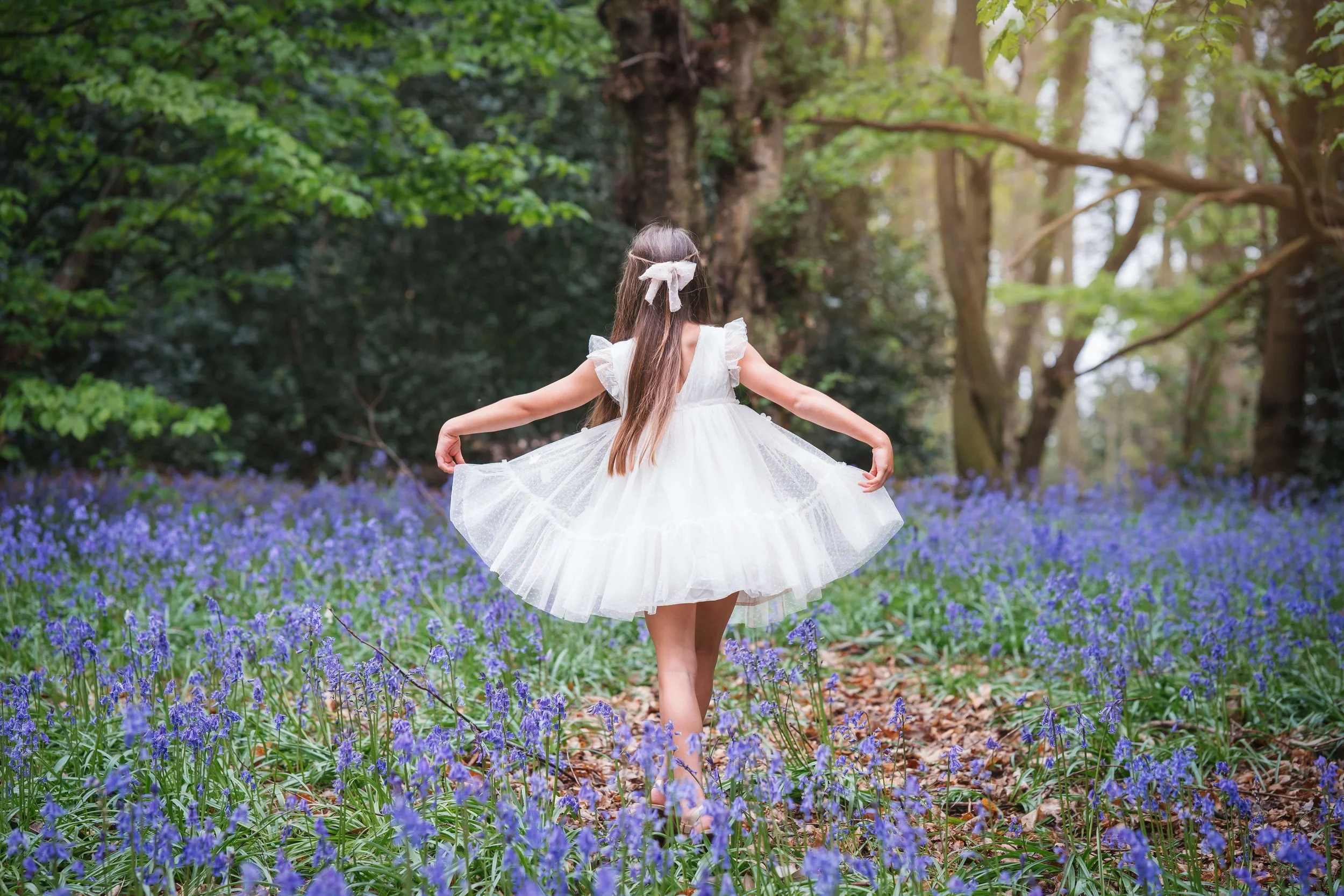 A girl in a white dress walking through a field of purple flowers in a forest during daylight.
