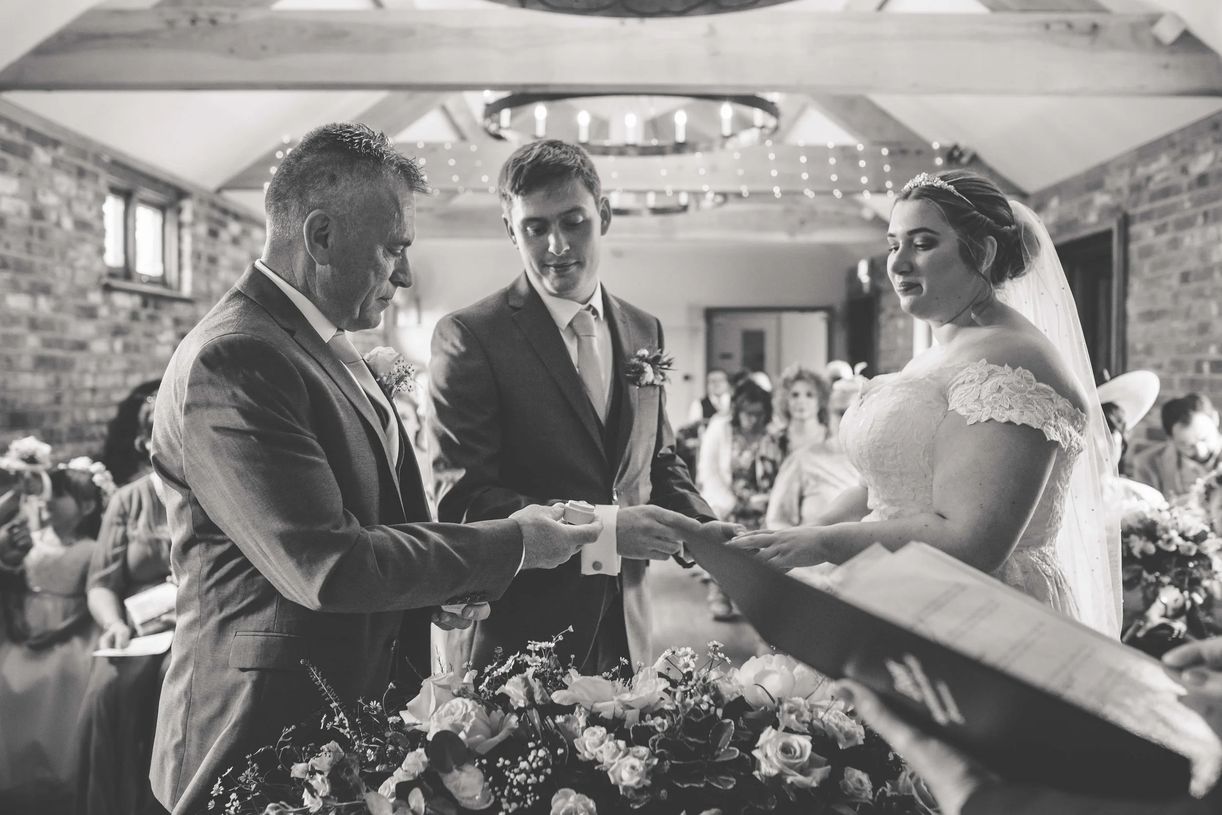 A black and white photo of a wedding ceremony showing a groom, bride, and officiant at the altar, with guests in the background.