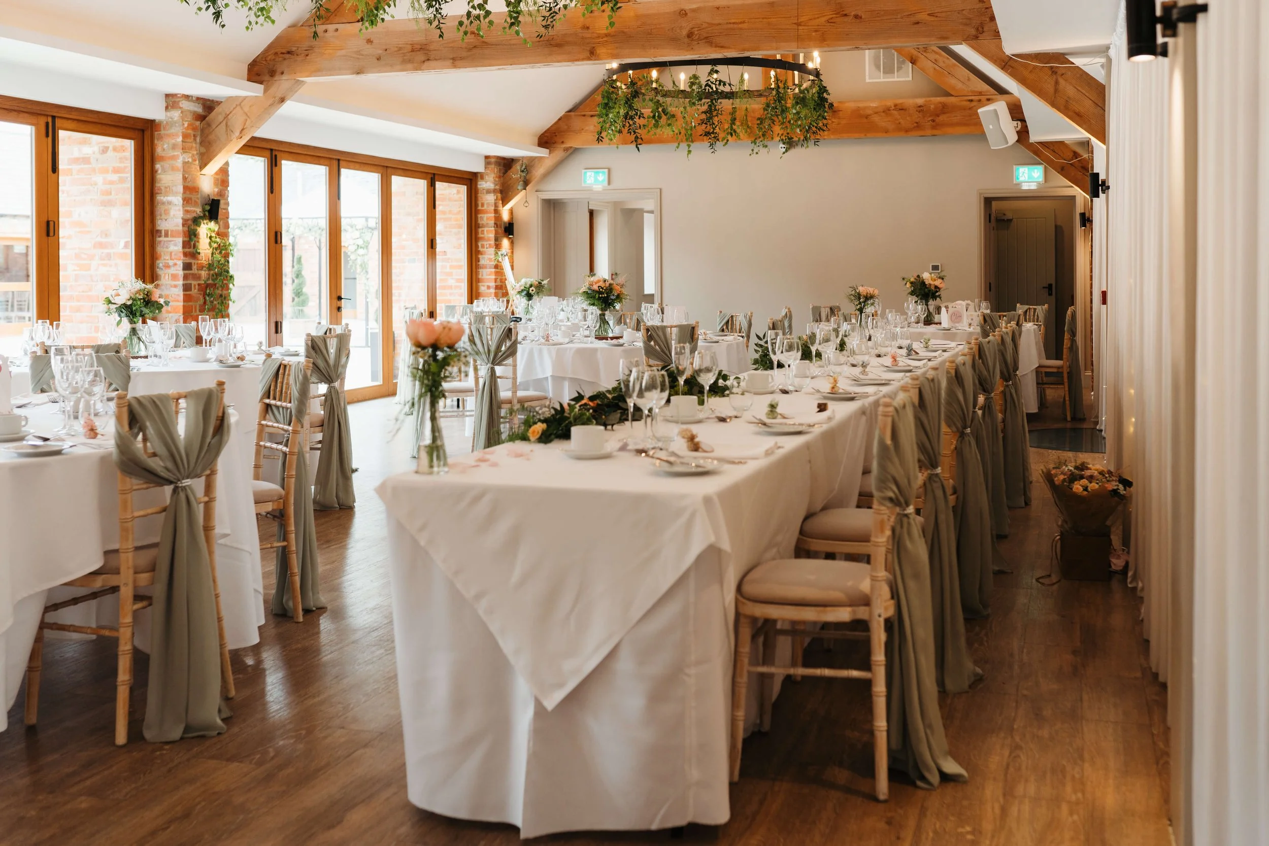Elegant wedding reception setup with long tables covered in white tablecloths, decorated with floral centerpieces, glassware, and china. Chairs have light fabric sashes, and the room features wooden beams, large windows, and brick walls with greenery