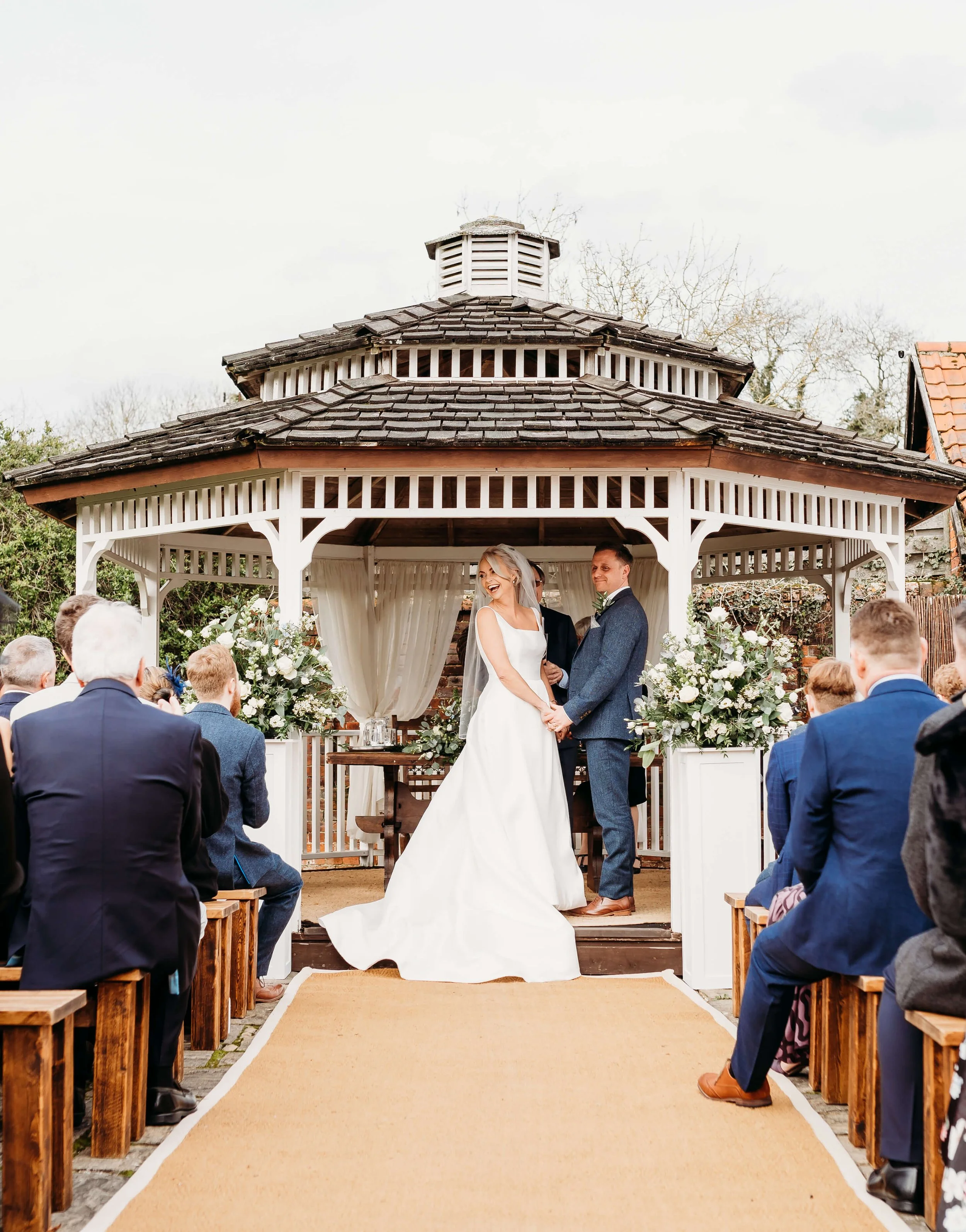 A couple gets married under a gazebo in an outdoor wedding ceremony, surrounded by seated guests. The bride and groom hold hands in front of floral arrangements, with the bride in a white gown and veil, and the groom in a blue suit.