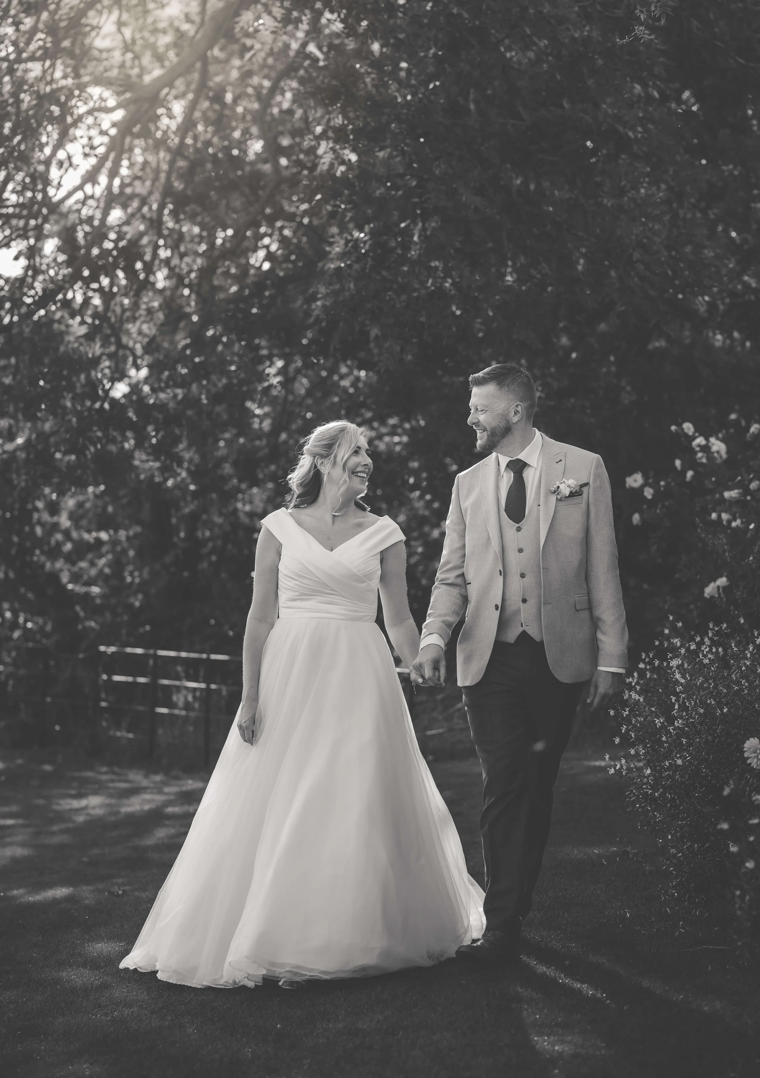 A black and white photo of a bride and groom walking hand in hand outdoors, smiling at each other, with trees and bushes in the background.