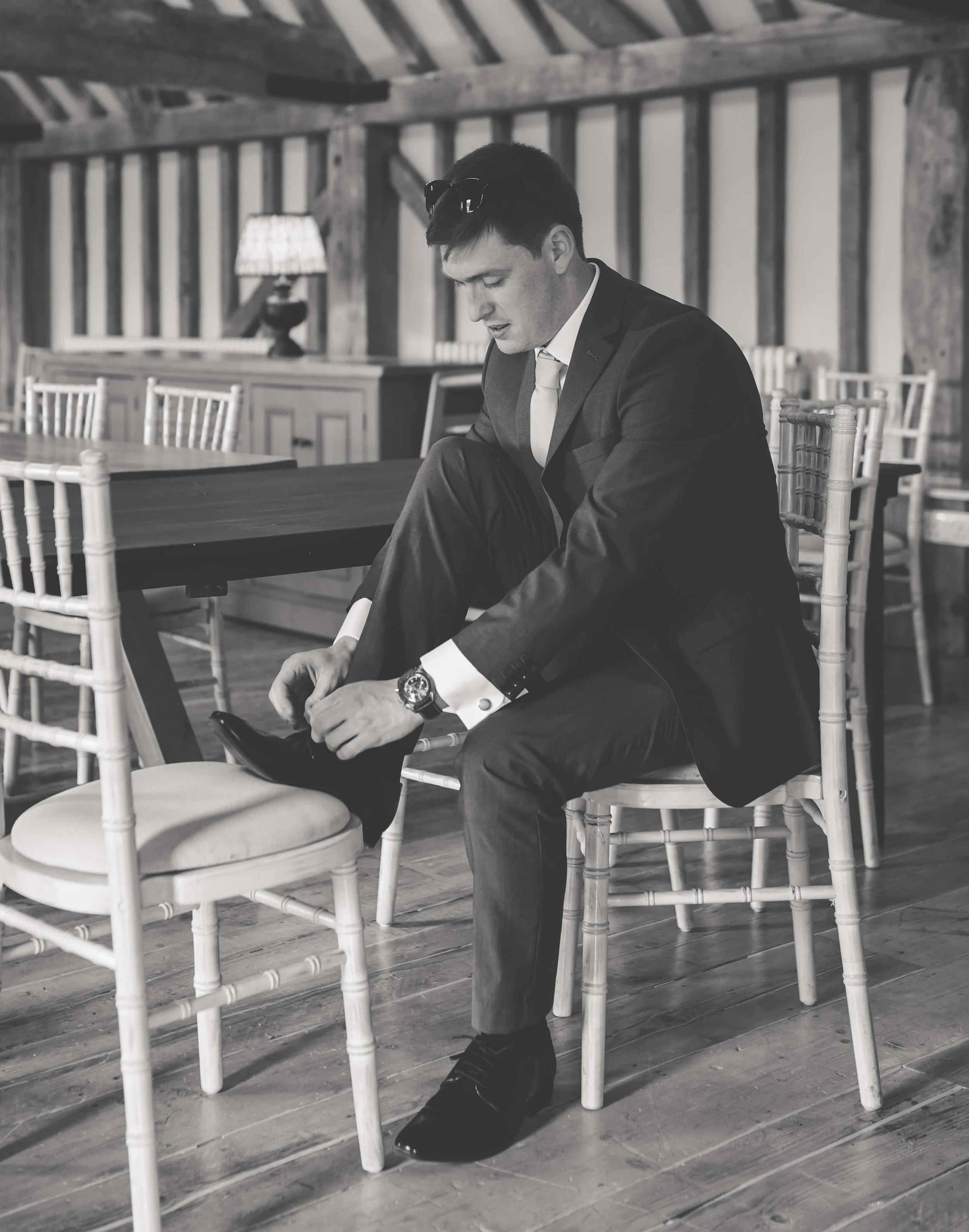 A man in a suit sitting on a chair, adjusting his shoe, in a rustic wooden room.