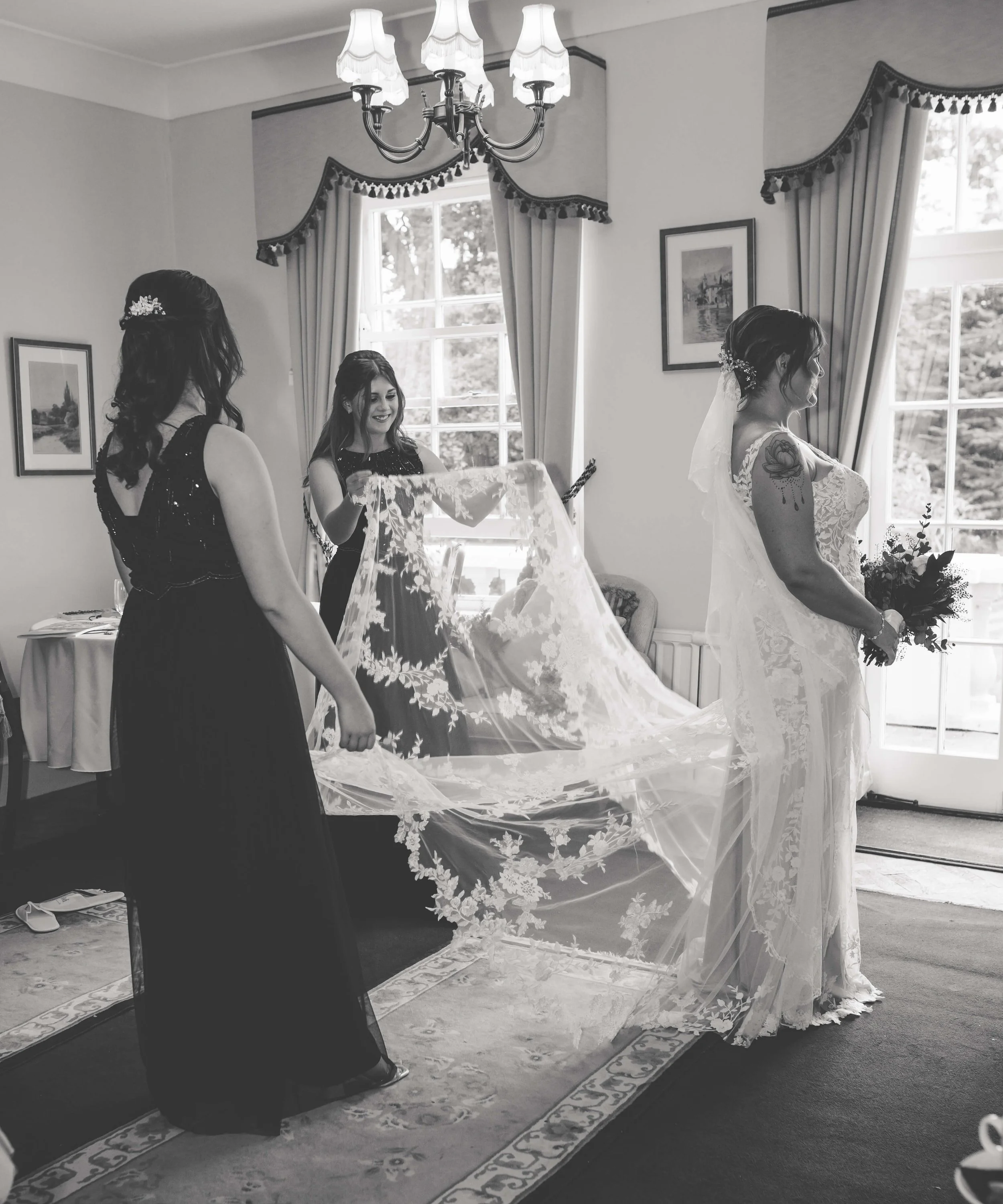 Black and white photo of a bride and two women in a room with large windows, curtains, and framed pictures, as the women help the bride with her veil.