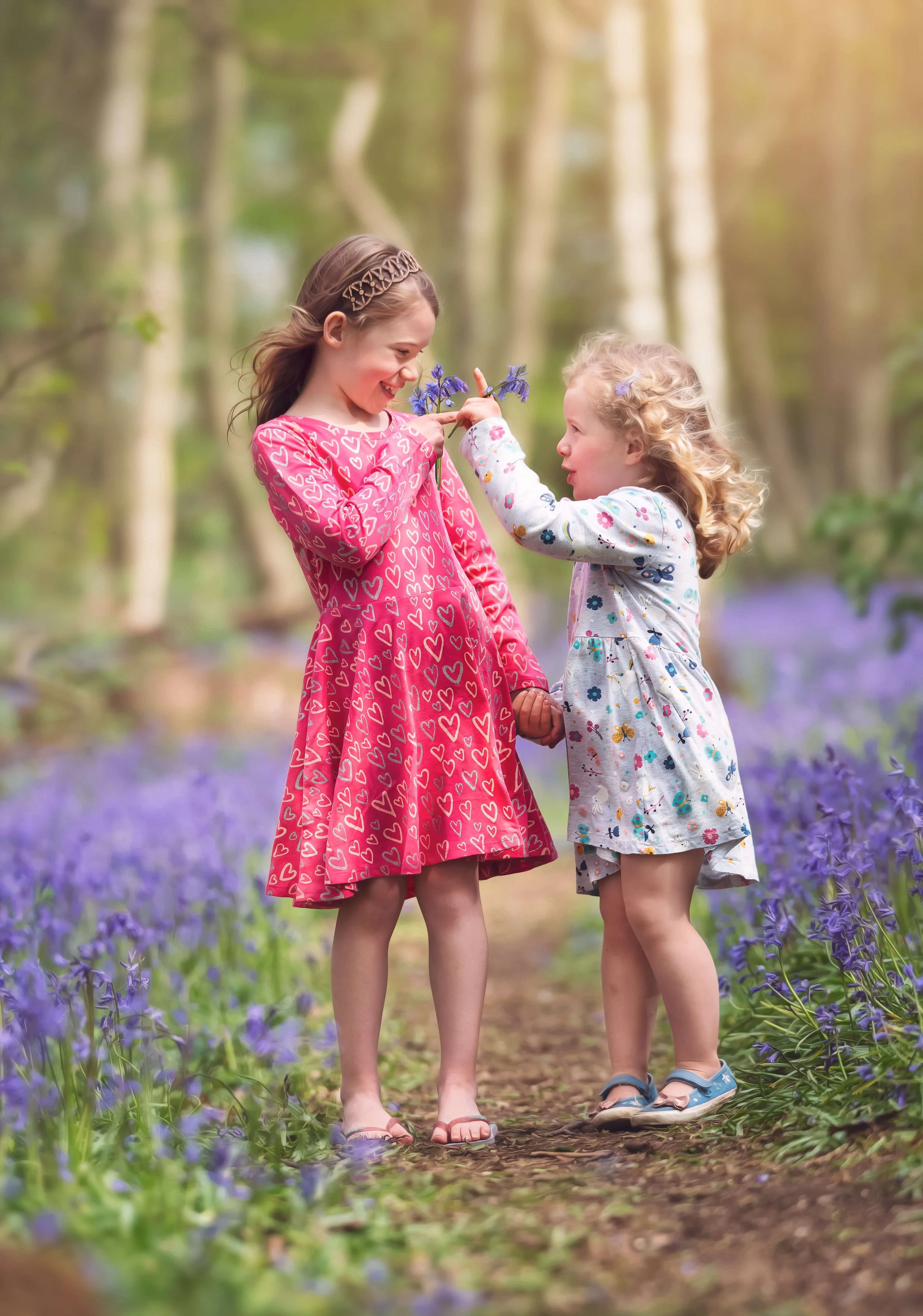 Two young girls are standing on a path in a wooded area, with purple flowers on either side. One girl with brown hair is smiling and holding a bunch of purple flowers, while the other girl with blonde curly hair is touching the flowers to the first g