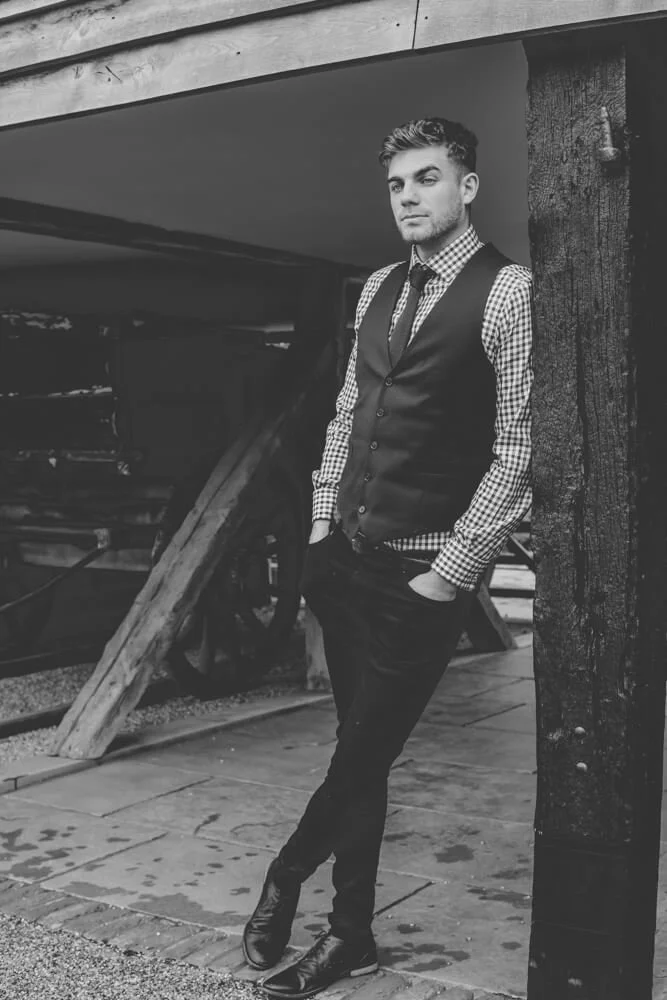A young man with styled hair wearing a checkered shirt, vest, and tie, standing outdoors next to a wooden post, with hands in pockets, looking off to the side. Black and white photograph.