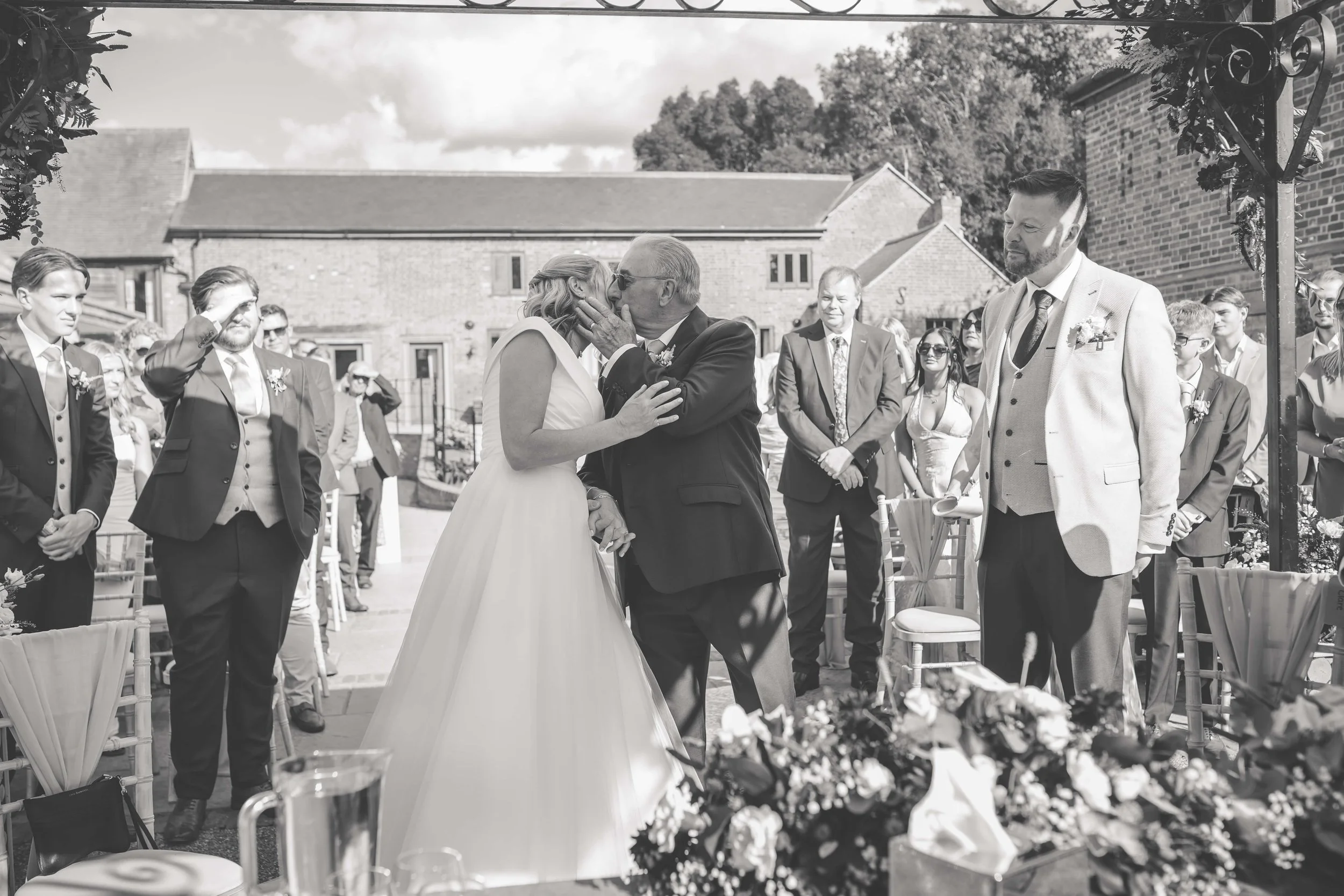 A wedding ceremony outdoors with the bride and her father sharing a kiss while the groom looks on. Guests are standing around, some saluting, on a sunny day with a backdrop of brick buildings.