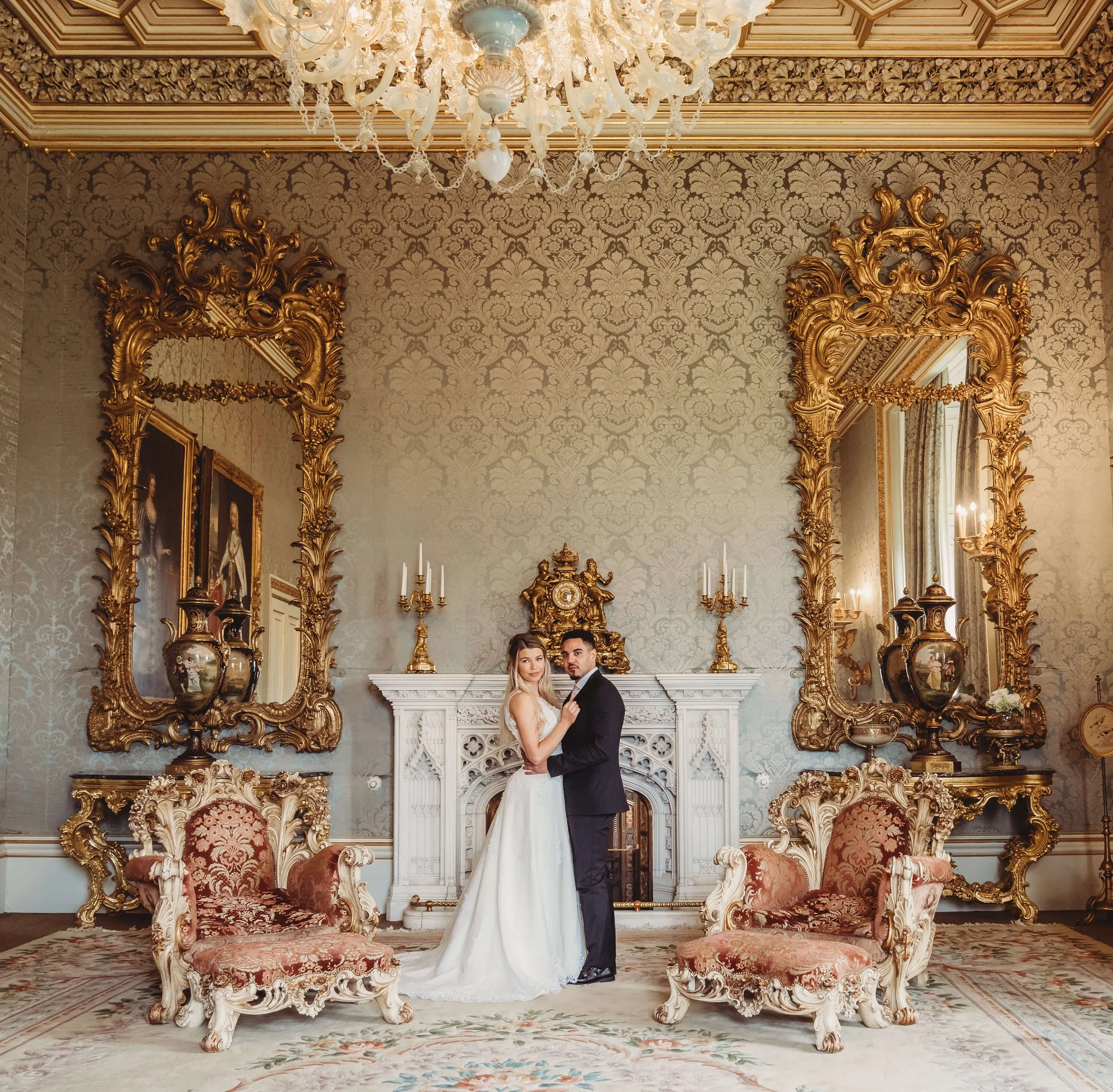 A couple in wedding attire standing in an ornate, luxurious room with gold-framed mirrors, antique furniture, and elaborate decorations.
