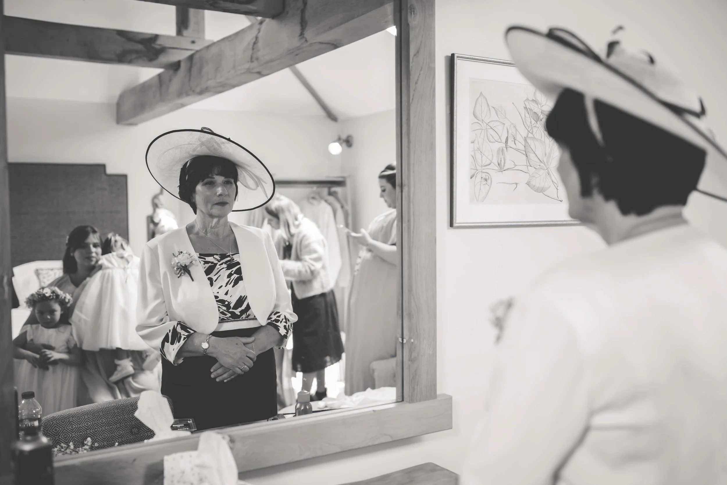 A woman wearing a large sunhat and a floral dress looks at herself in a mirror, surrounded by women and children in a dressing room preparing for a formal event.
