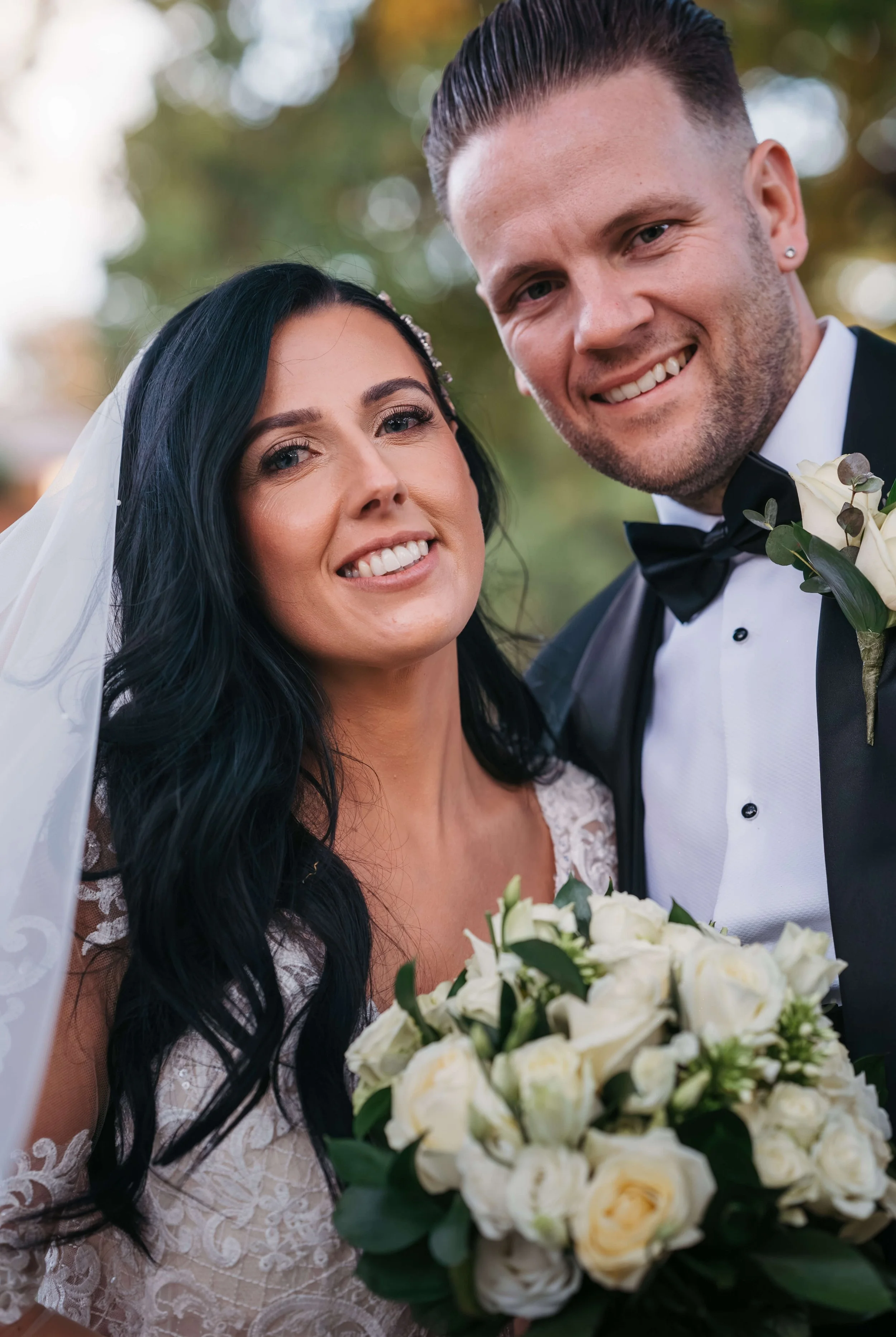 A bride and groom smiling outdoors, the bride holding a bouquet of white roses and greenery, both dressed in wedding attire.
