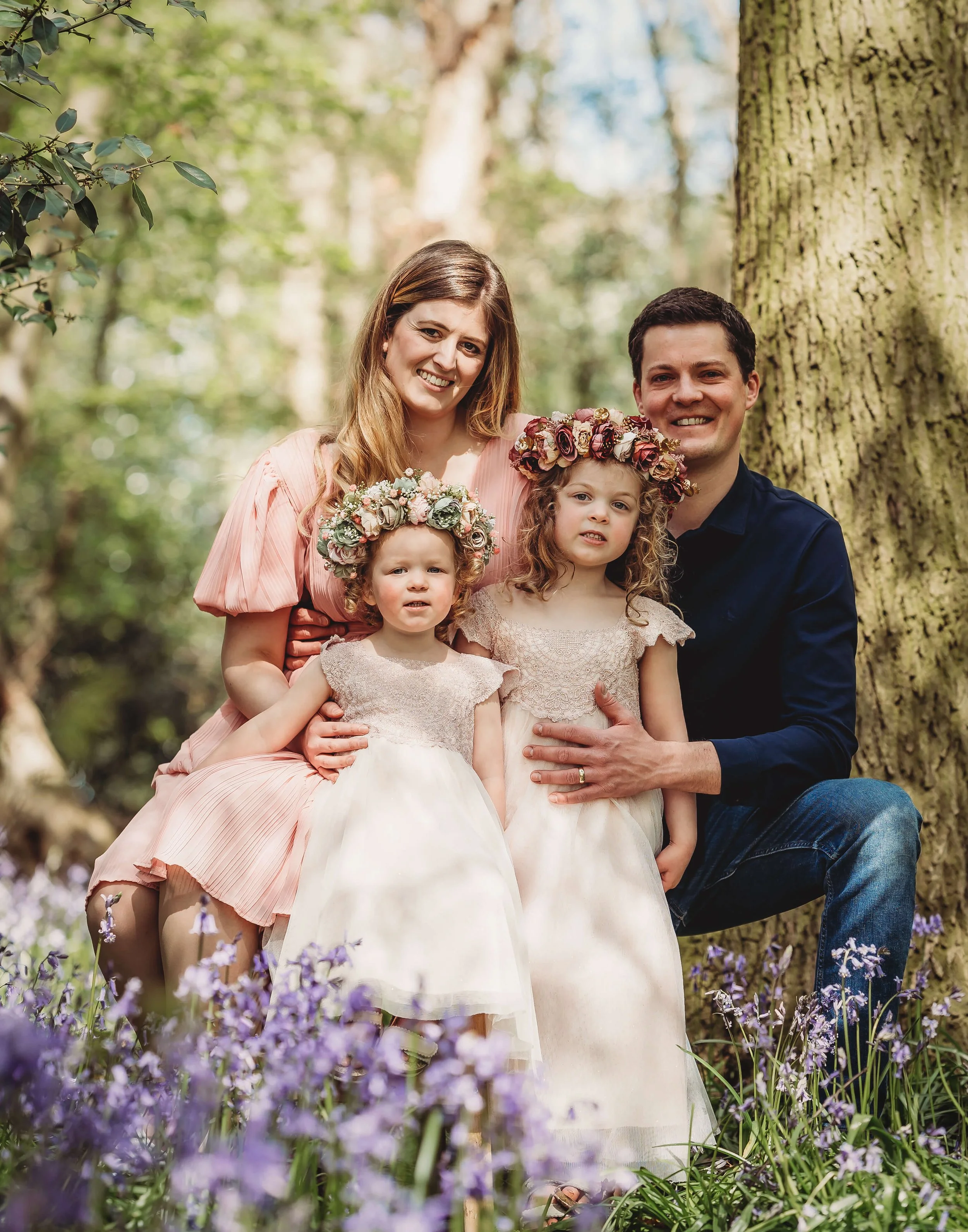 A family of four outdoors in a wooded area during spring or summer, with purple flowers in the foreground. The mother and father are smiling, with the mother wearing a pink dress and the father in a dark shirt. The two young girls are dressed in whit