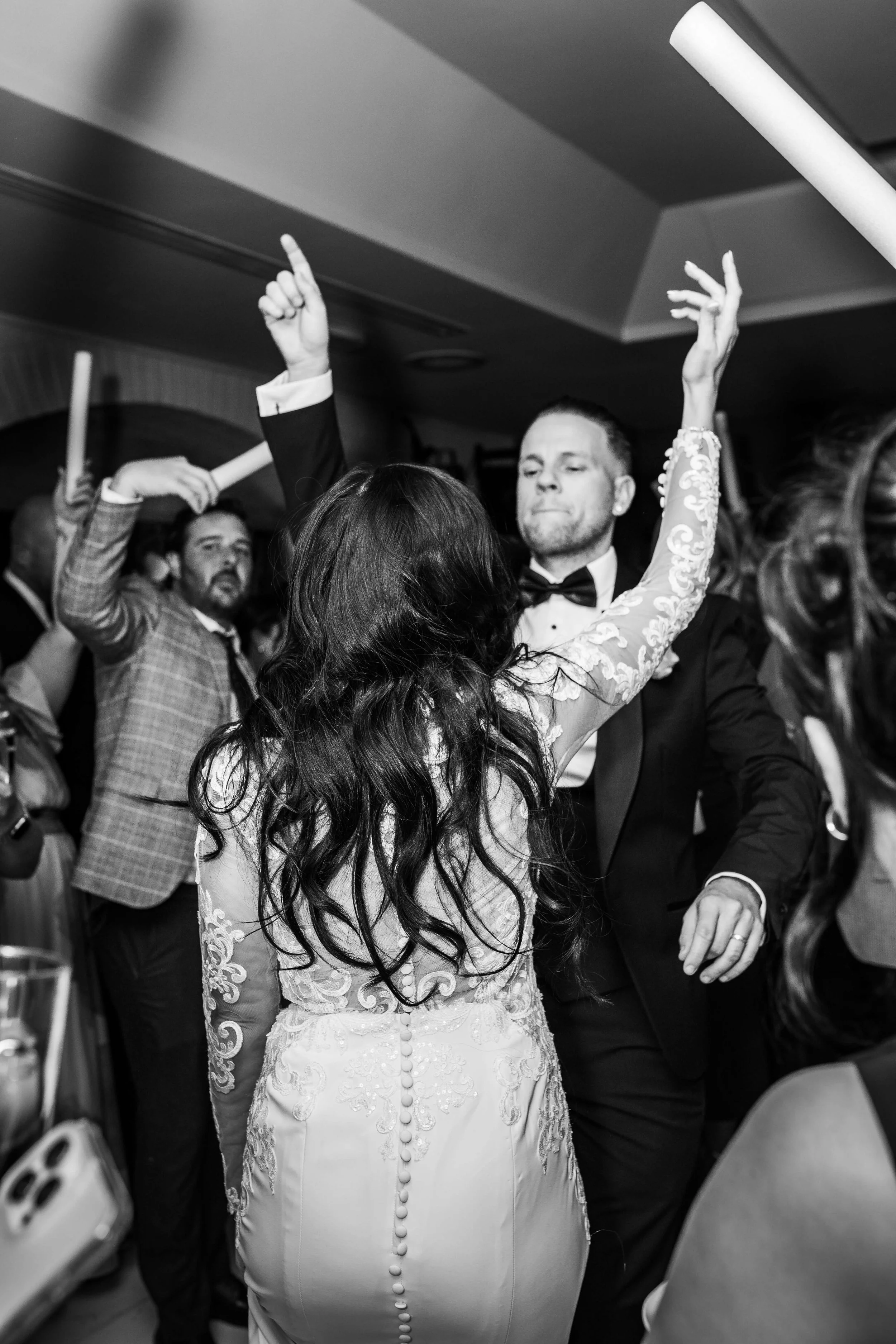 A black and white photo of a wedding reception dance floor with guests dancing. In the foreground, a woman with long dark hair in a wedding gown is dancing with a man in a tuxedo. Other guests are visible in the background, also dancing.
