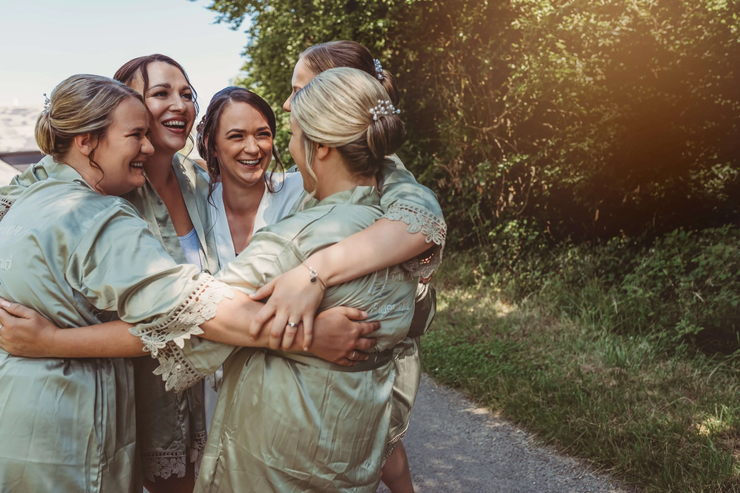 Group of women in matching satin robes hugging and smiling outdoors on a sunny day.