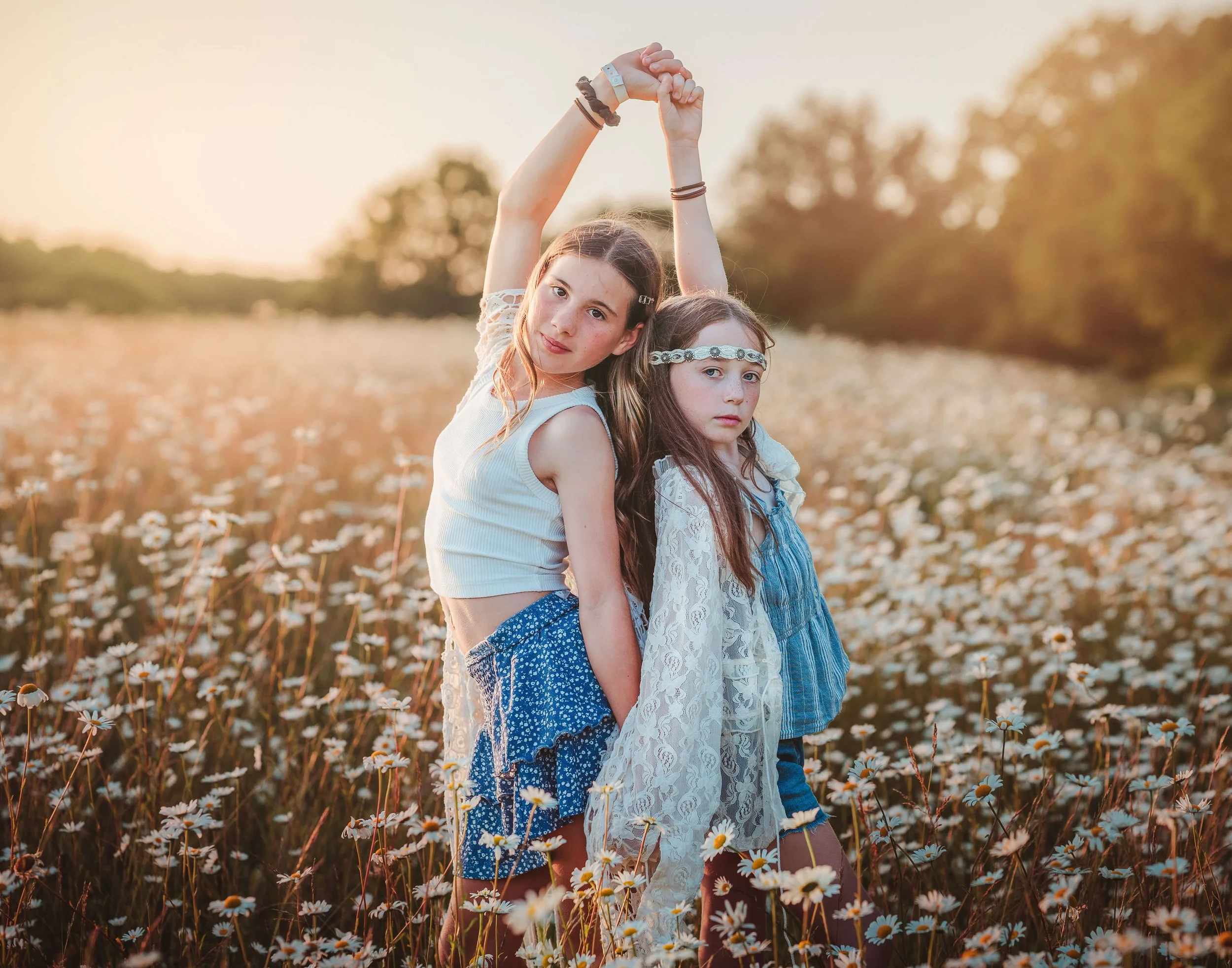 Two young girls standing in a field of daisies during sunset, holding hands raised above their heads.