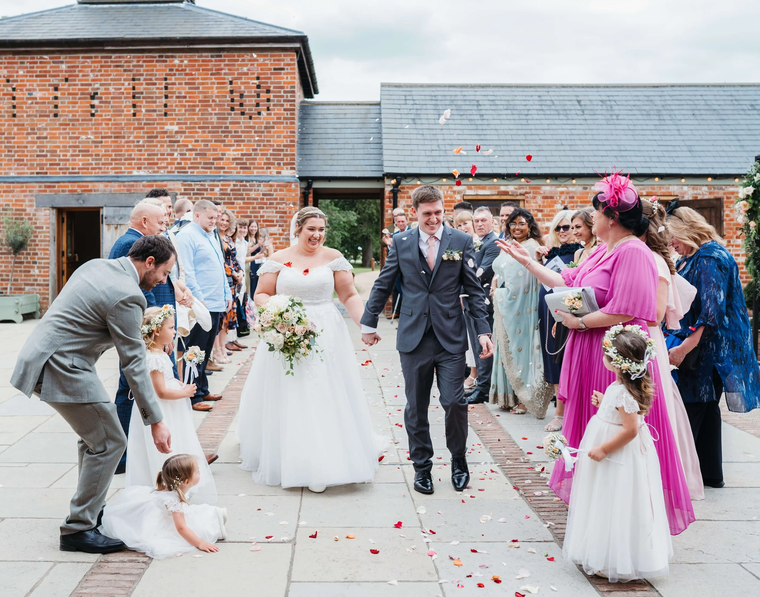 Bride and groom walking hand-in-hand down a flower-strewn aisle outside a brick building, surrounded by friends and family celebrating their wedding with flower petals in the air.