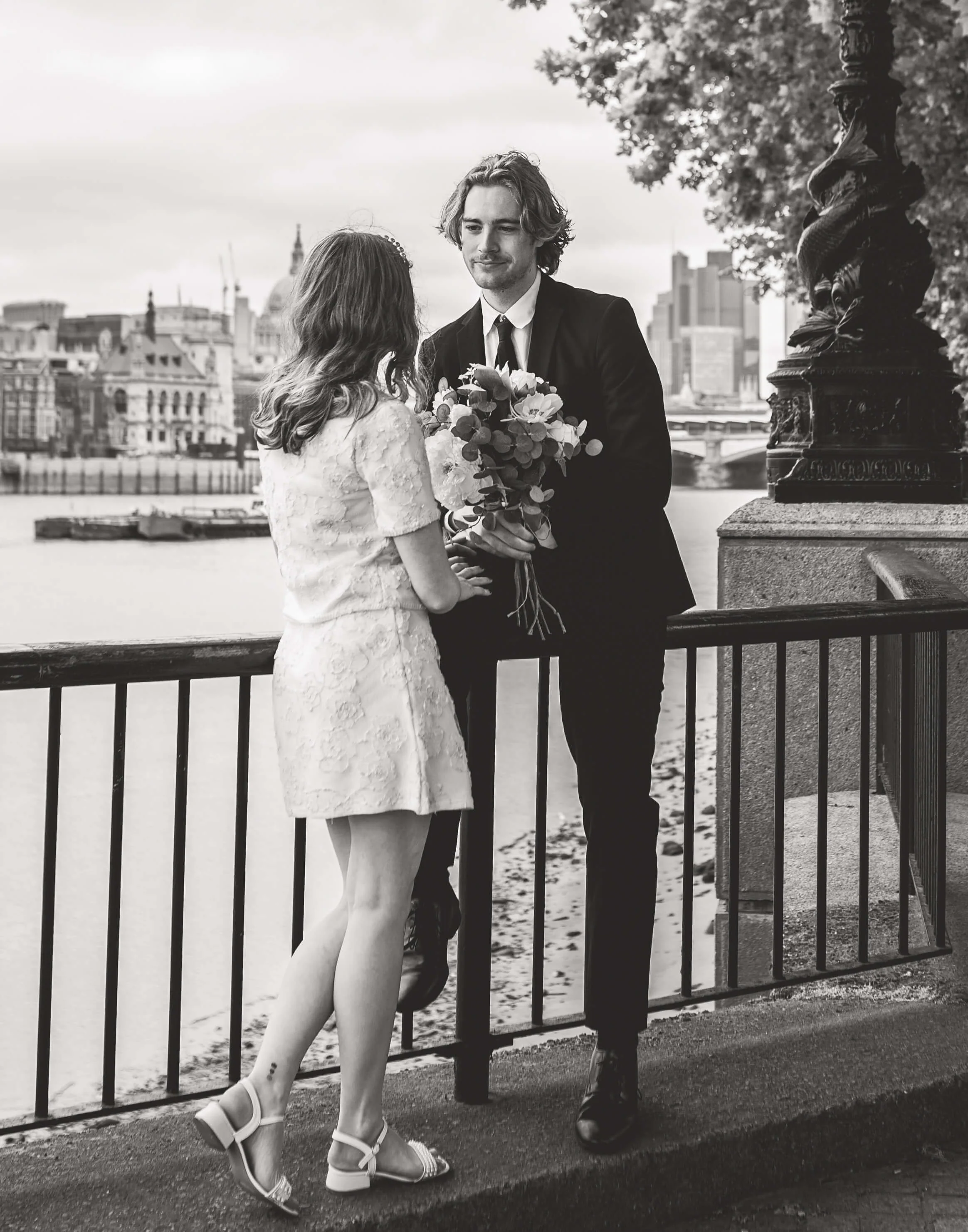 A black and white photo of a romantic moment between a young woman in a lace dress and a young man in a suit, with the woman holding a bouquet of flowers, standing on a riverside with city buildings in the background.