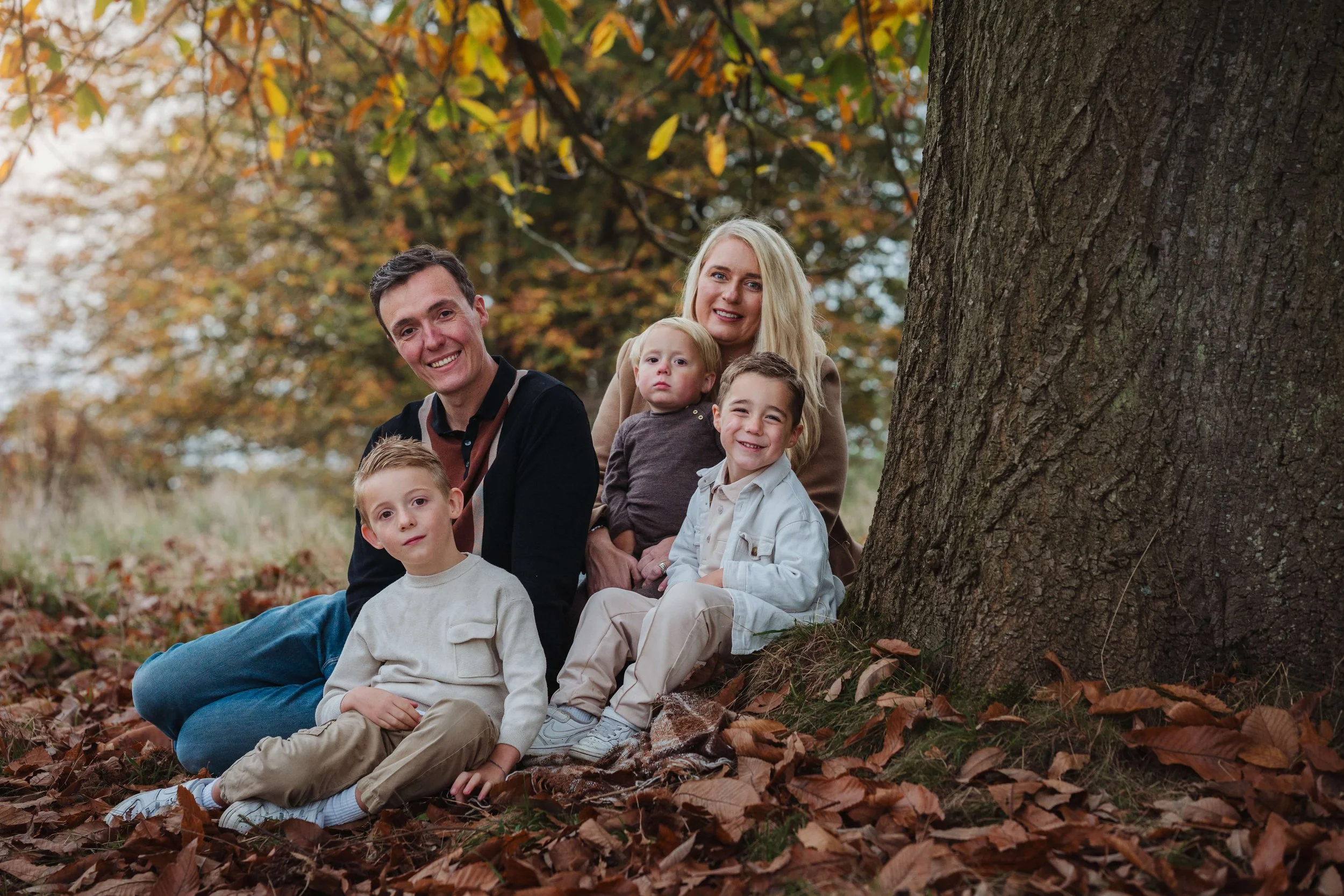 A family of six sitting on autumn leaves near a large tree in a park during fall, smiling and posing for the photo.