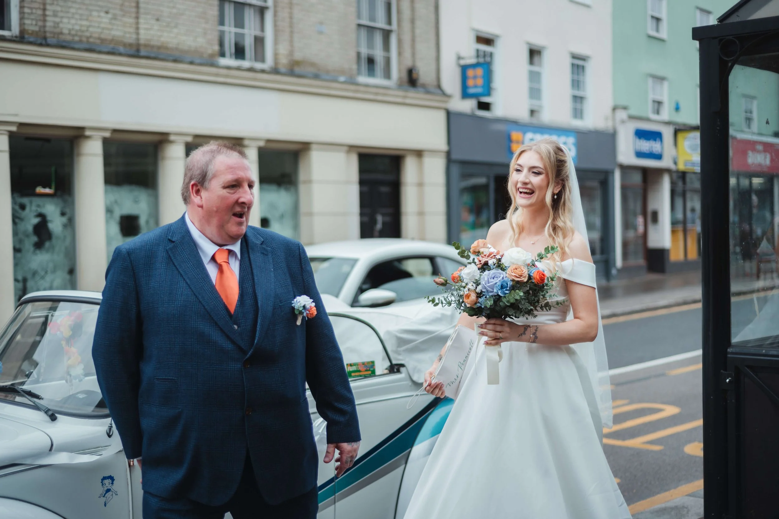 A bride in a white wedding dress holding a bouquet of flowers, smiling, and talking to a man in a blue suit with an orange tie on a city street.