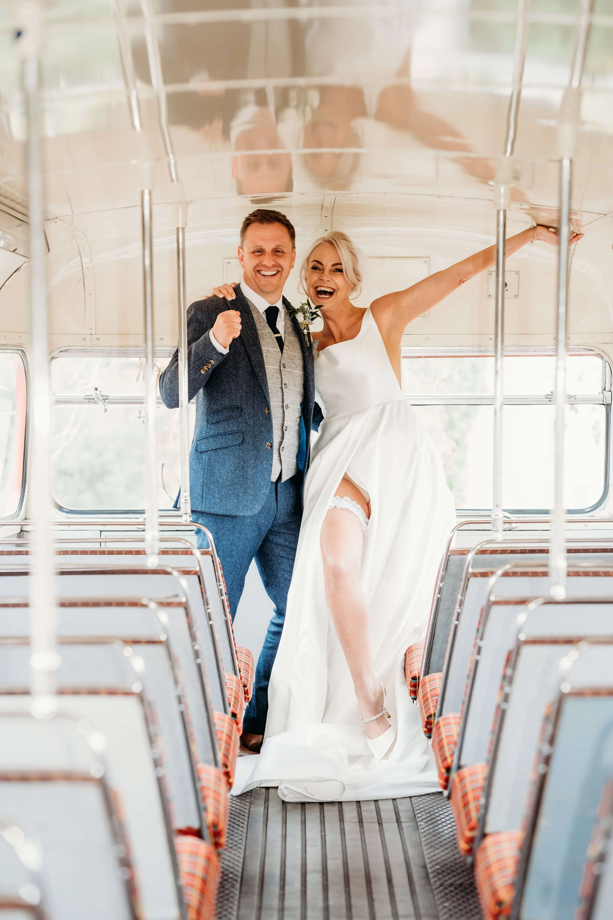 A bride and groom celebrating inside a vintage bus, with the bride lifting her leg and showing a garter, both smiling and dressed in wedding attire.
