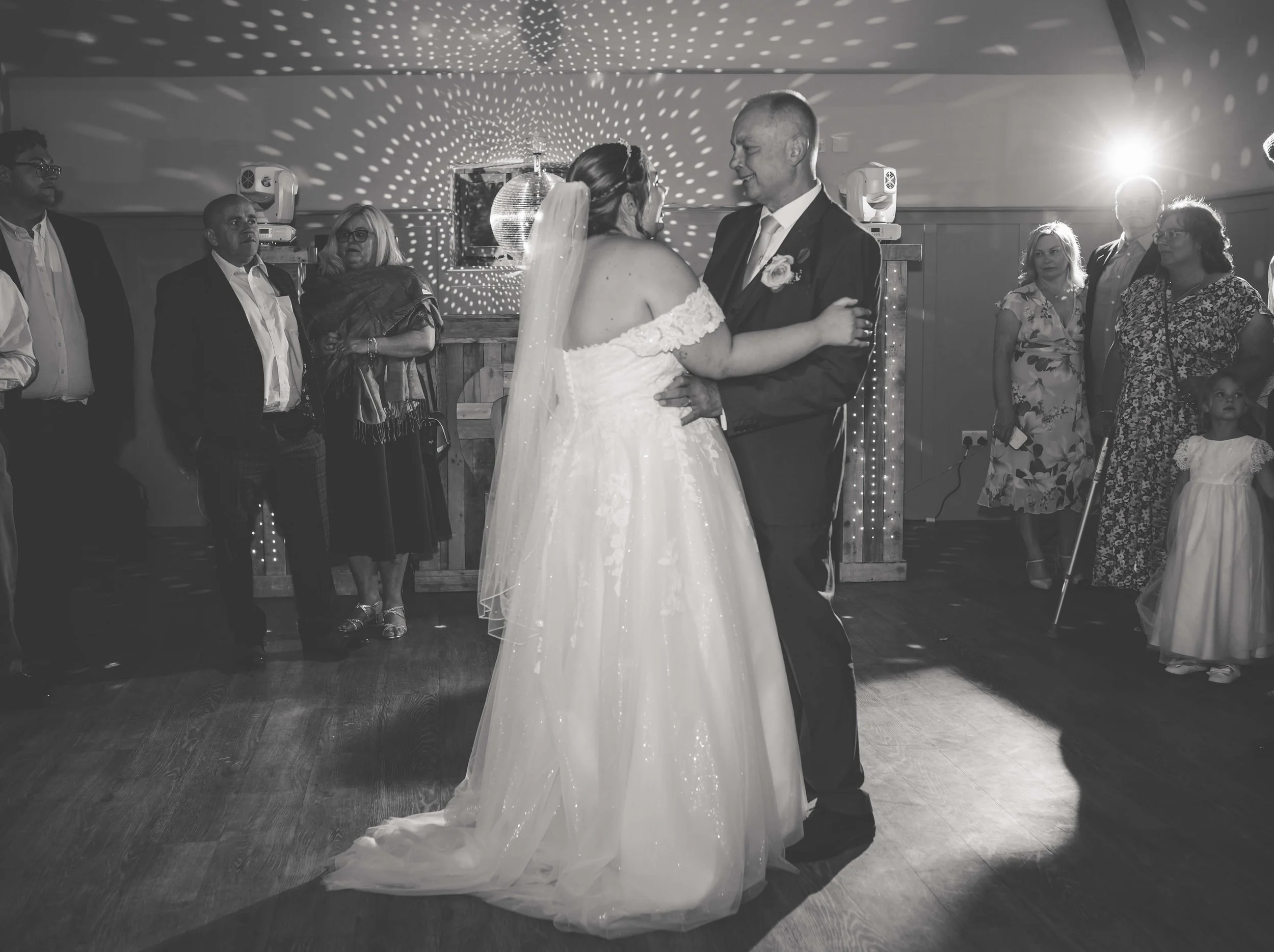 A bride and groom dancing at their wedding reception, surrounded by guests in a decorated venue with lights and a disco ball.